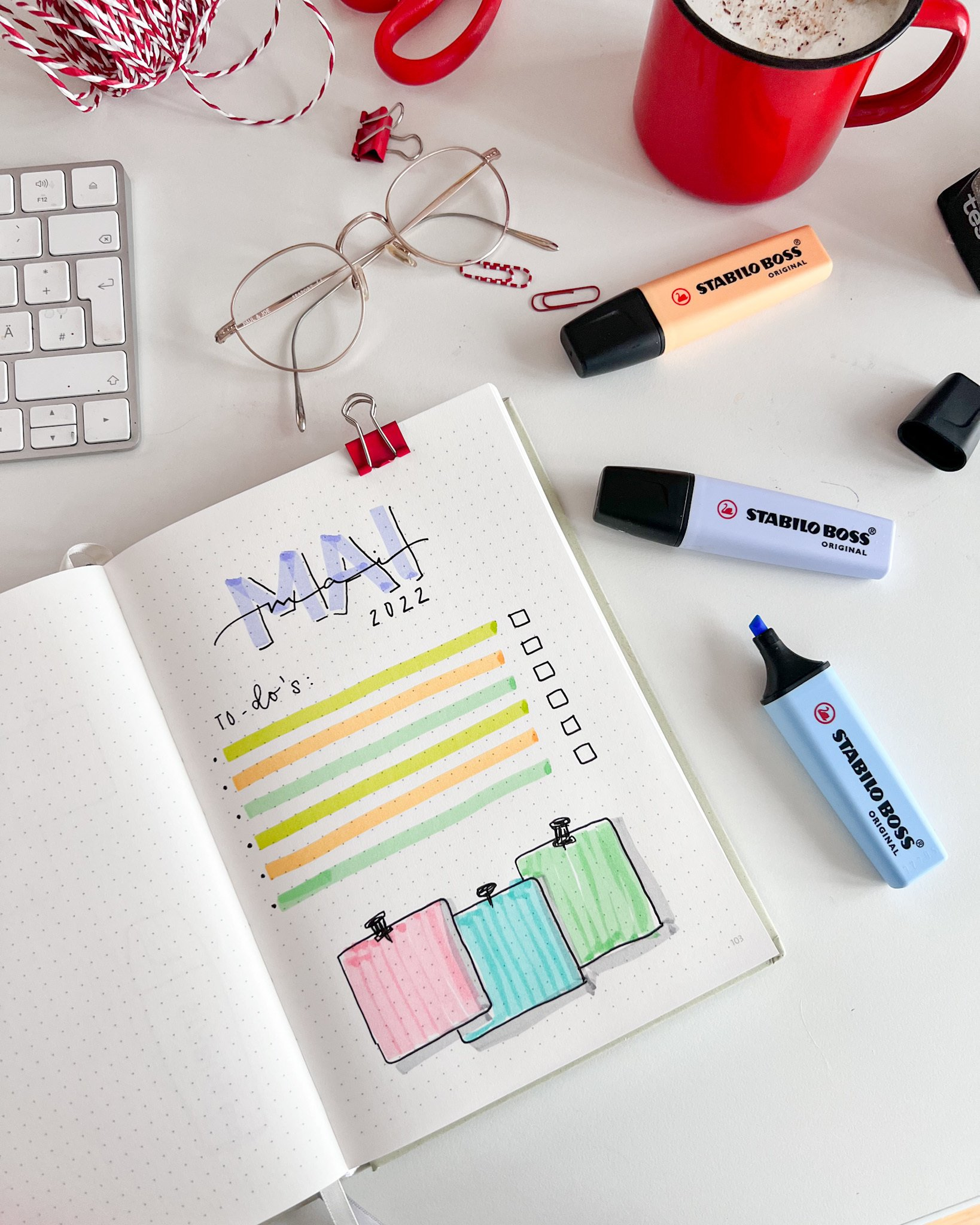 Ein Schreibtisch mit Notizbuch, Textmarkern, Brille, roter Tasse und Tastatur. Das Notizbuch ist zu einer To-Do-Liste geöffnet.