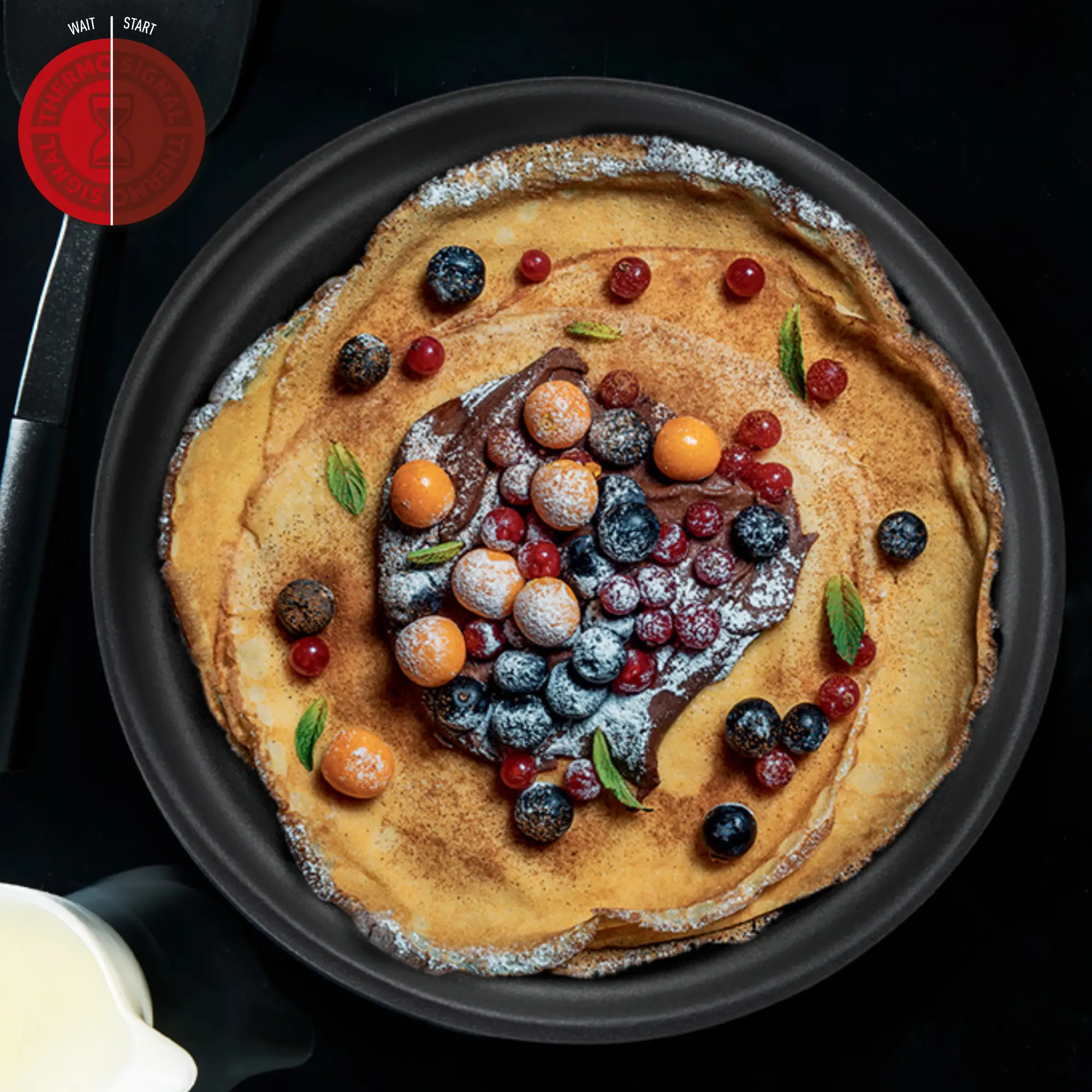 Ein Pfannkuchen auf einem schwarzen Teller, belegt mit Schokolade, Beeren und Minzblättern, mit Puderzucker bestäubt.