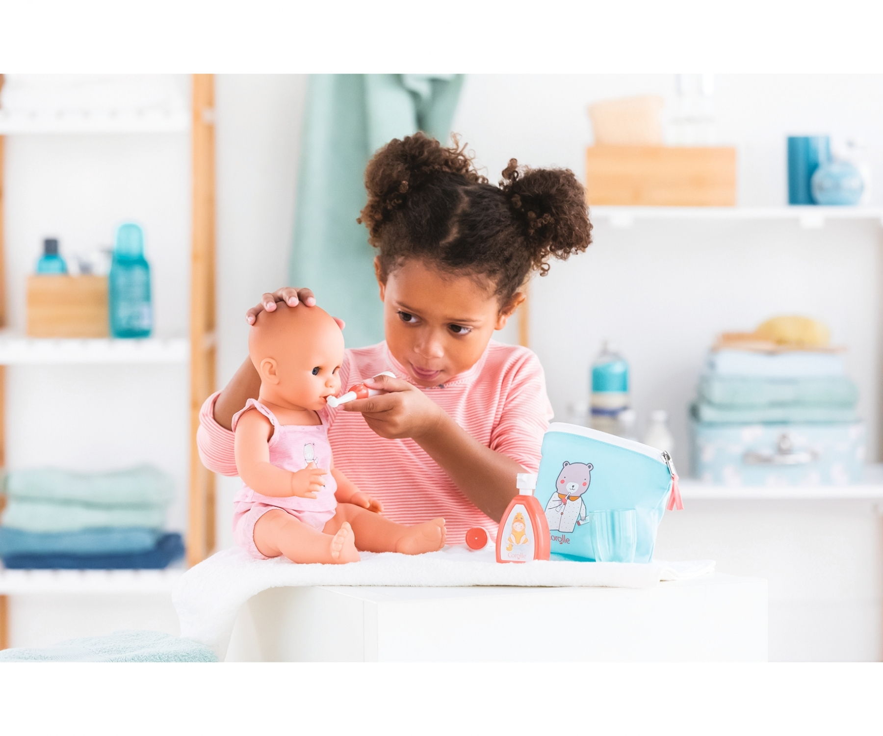 Dziewczynka szczotkuje zęby lalce. Niebieska torba, balsam i czerwona nakrętka leżą na białym stole.