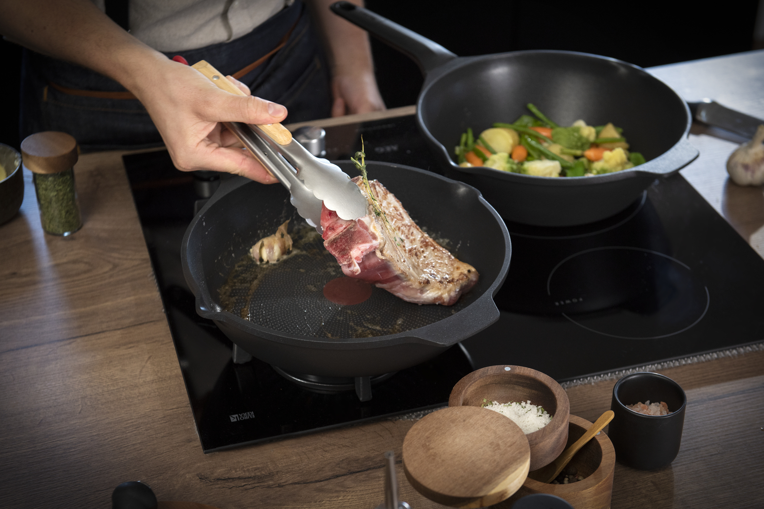 Een persoon draait met een tang een biefstuk in een pan. Groenten zitten in een andere pan. Kruiden en zout op tafel.
