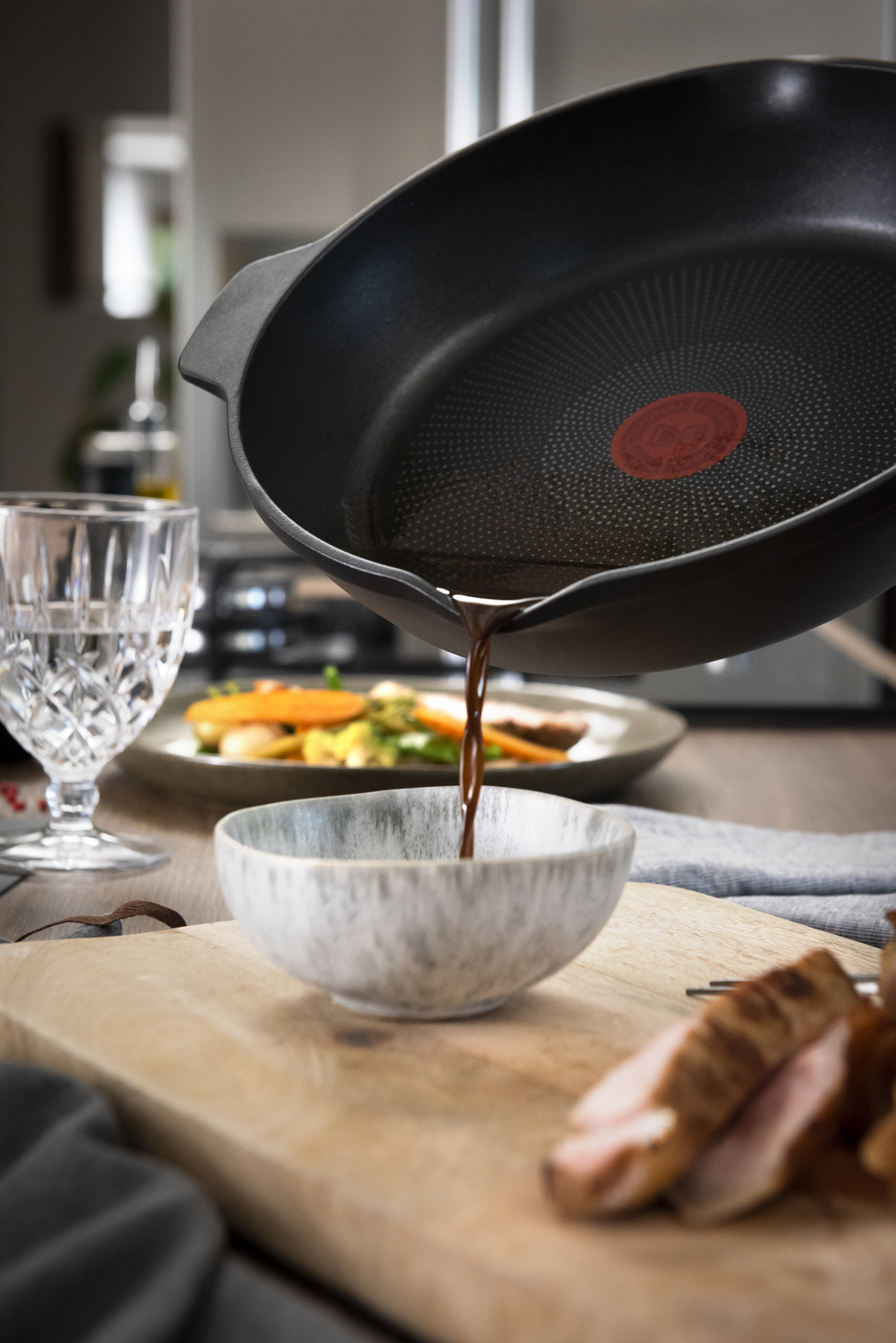 Saus wordt uit een pan in een kom gegoten. Eten en glas staan op tafel. De focus ligt op het schenken van de saus.