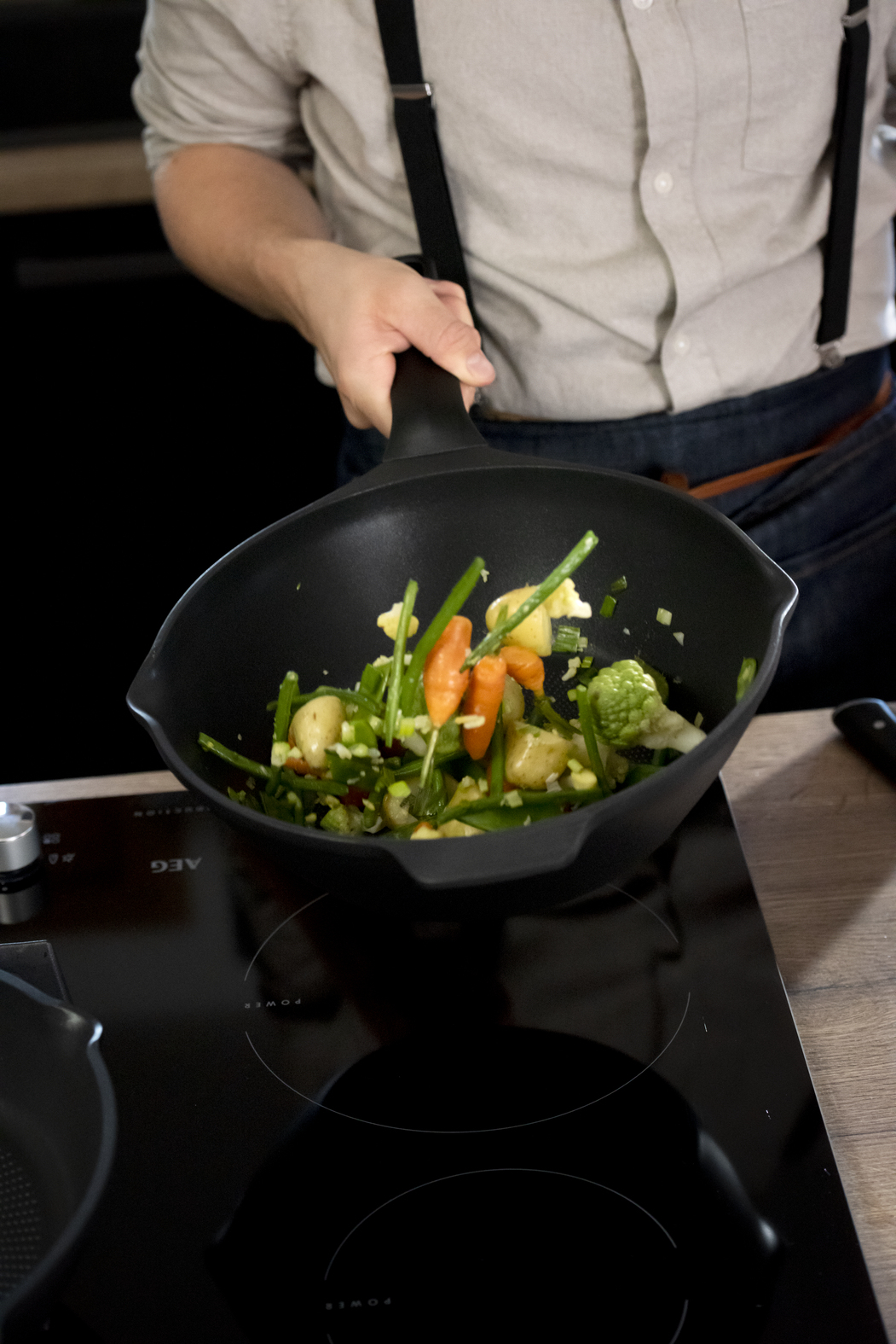Een persoon houdt een pan met groenten vast. Wortelen, broccoli en sperziebonen zijn zichtbaar. De pan staat op een zwart fornuis.