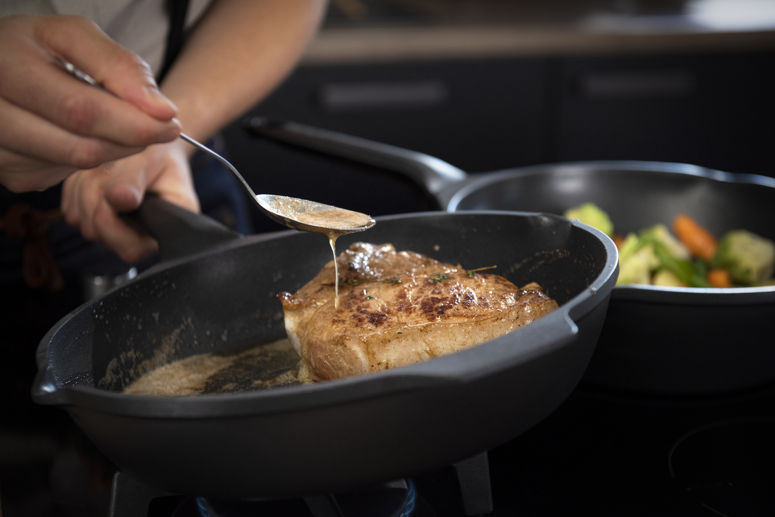 Een persoon giet saus over een gebakken biefstuk in een pan. Een andere pan met groenten op de achtergrond. Close-up.