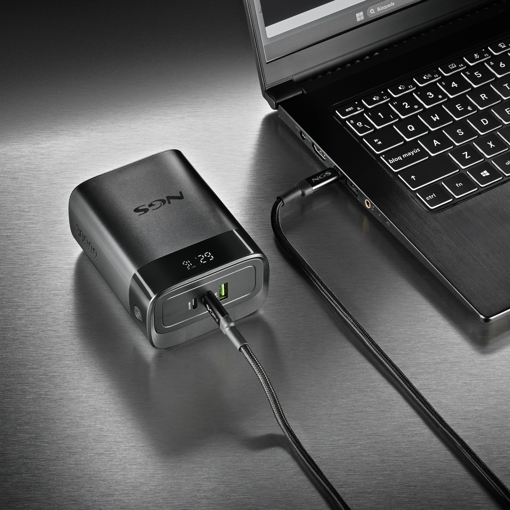 Banco de energía negro conectado a una laptop con un cable USB sobre una superficie metálica. El banco tiene pantalla.