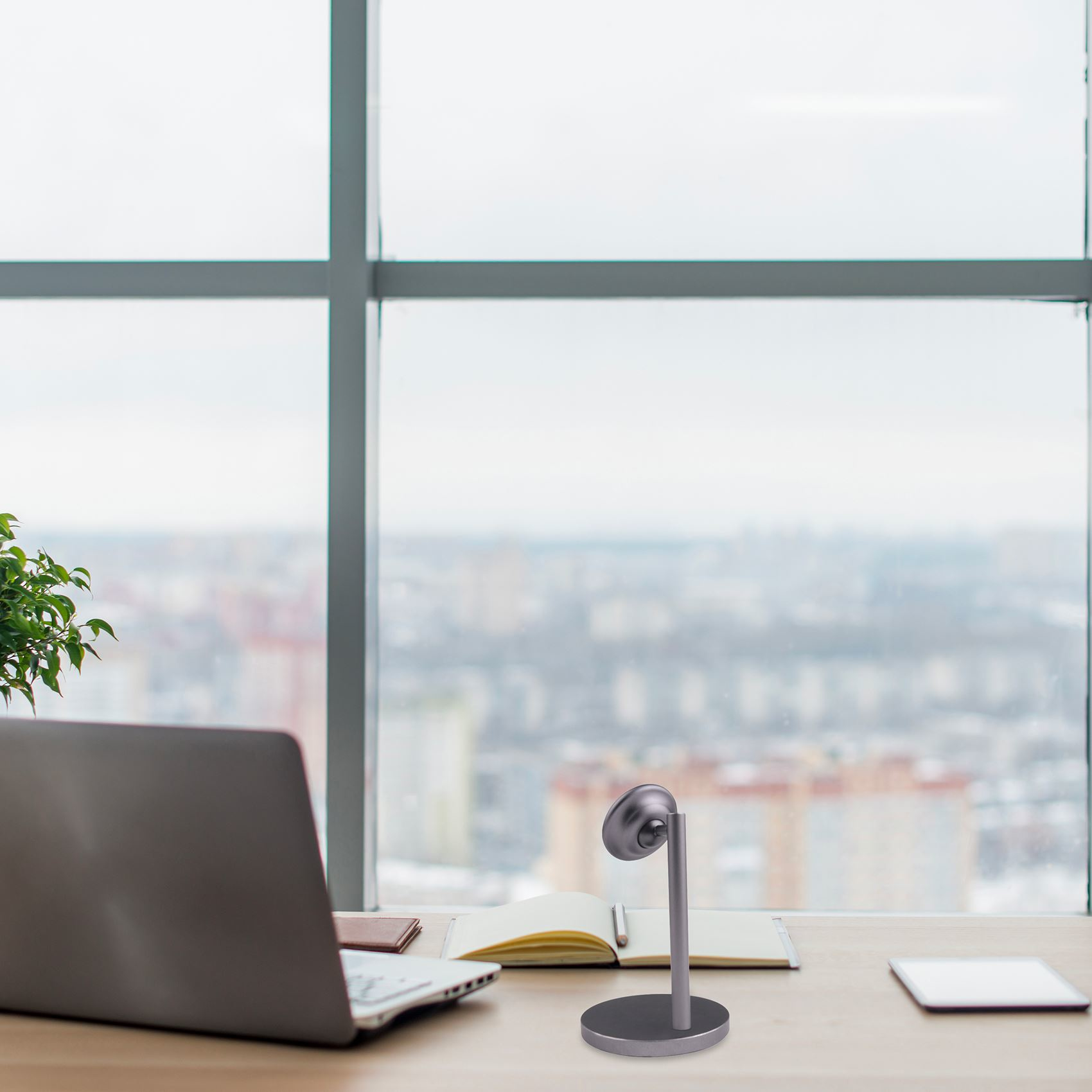 Ein Schreibtisch mit Laptop, Lampe, Notizblock und Tablet. Ein Fenster mit Blick auf die Stadt im Hintergrund. Eine Pflanze links.