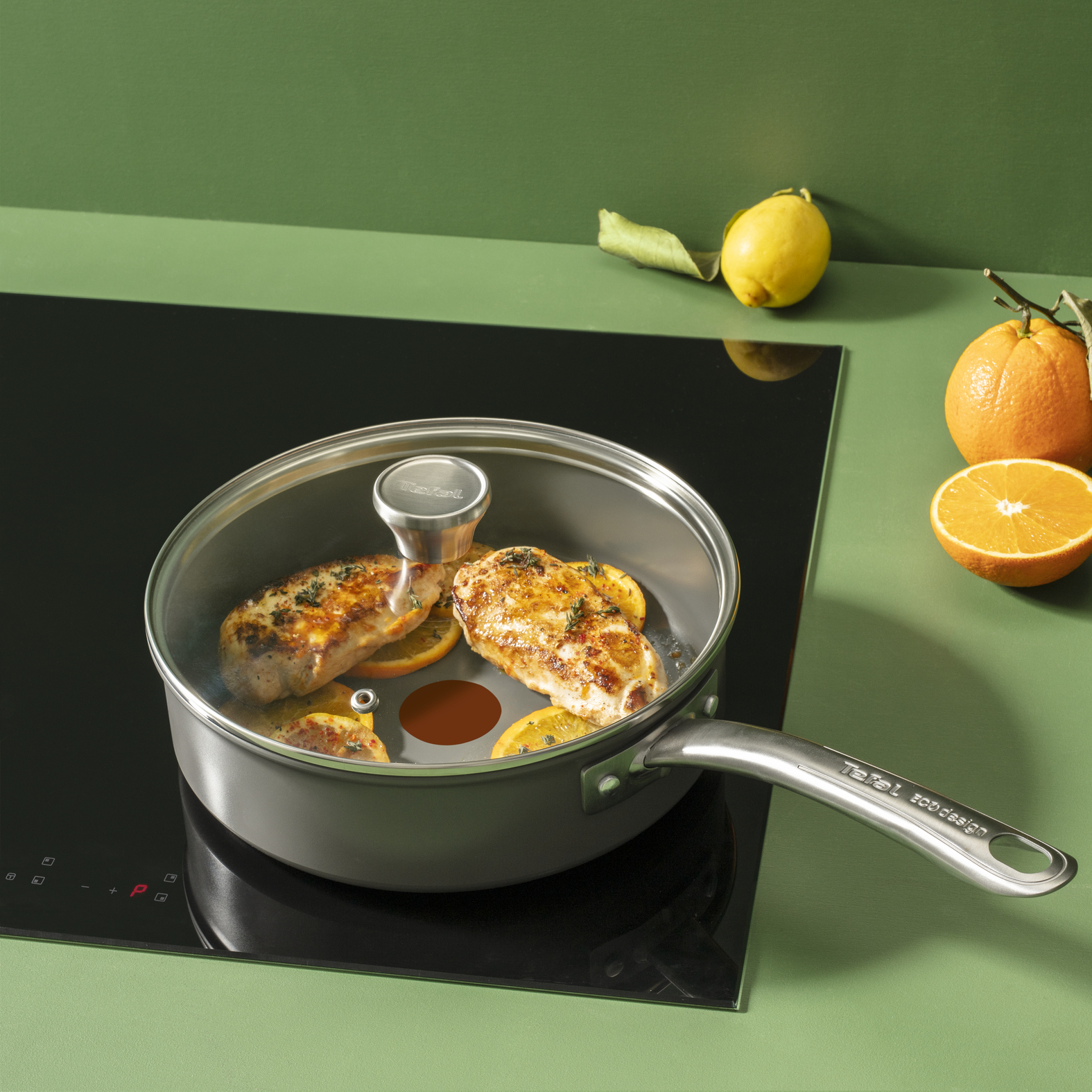 Hähnchenbrüste mit Orangenscheiben in der Pfanne. Zitrone und Orange daneben, auf grüner Fläche mit Induktionskochfeld.