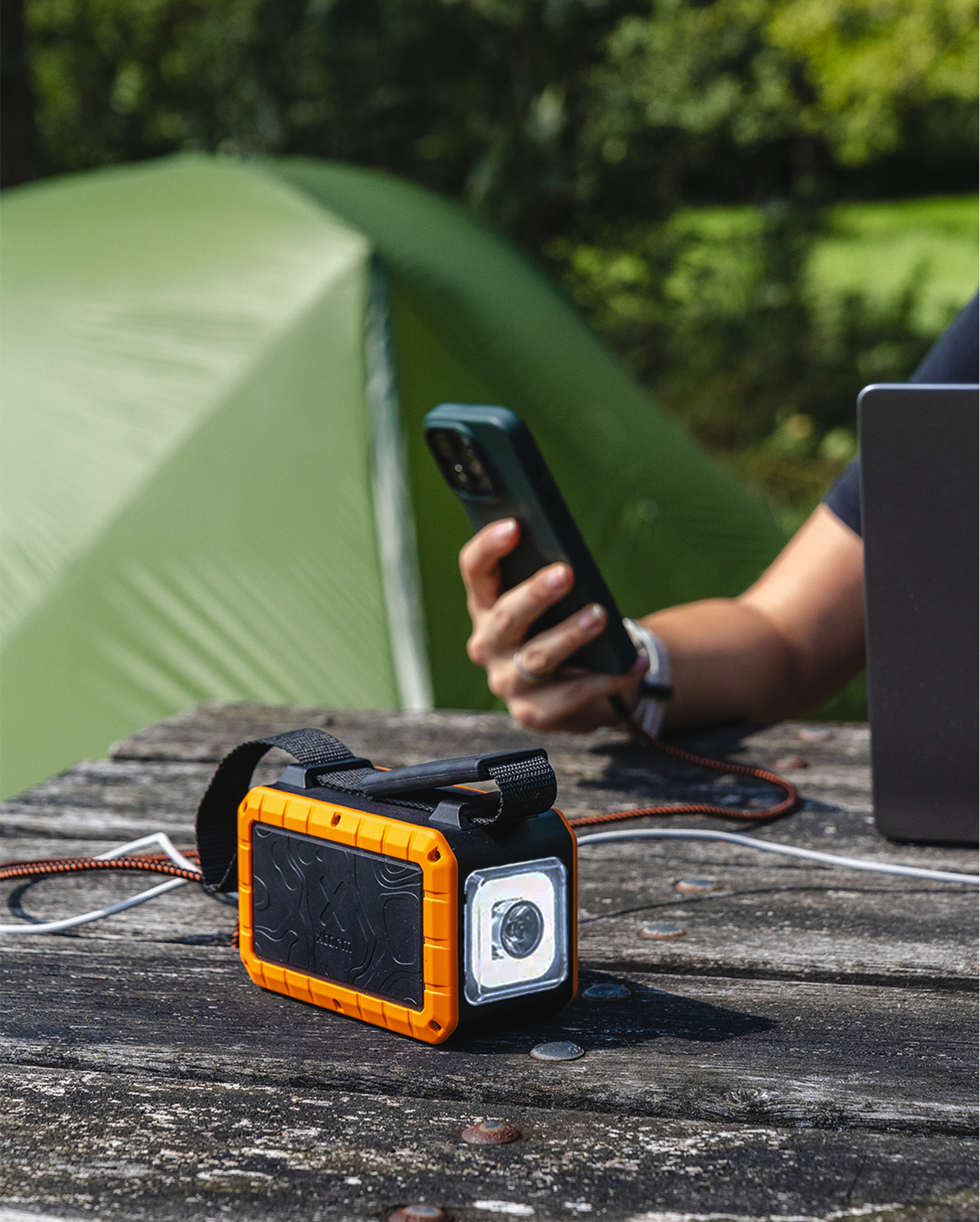 Une lampe de poche orange et noire sur une table en bois. Une personne tient un téléphone et une tente est en arrière-plan.