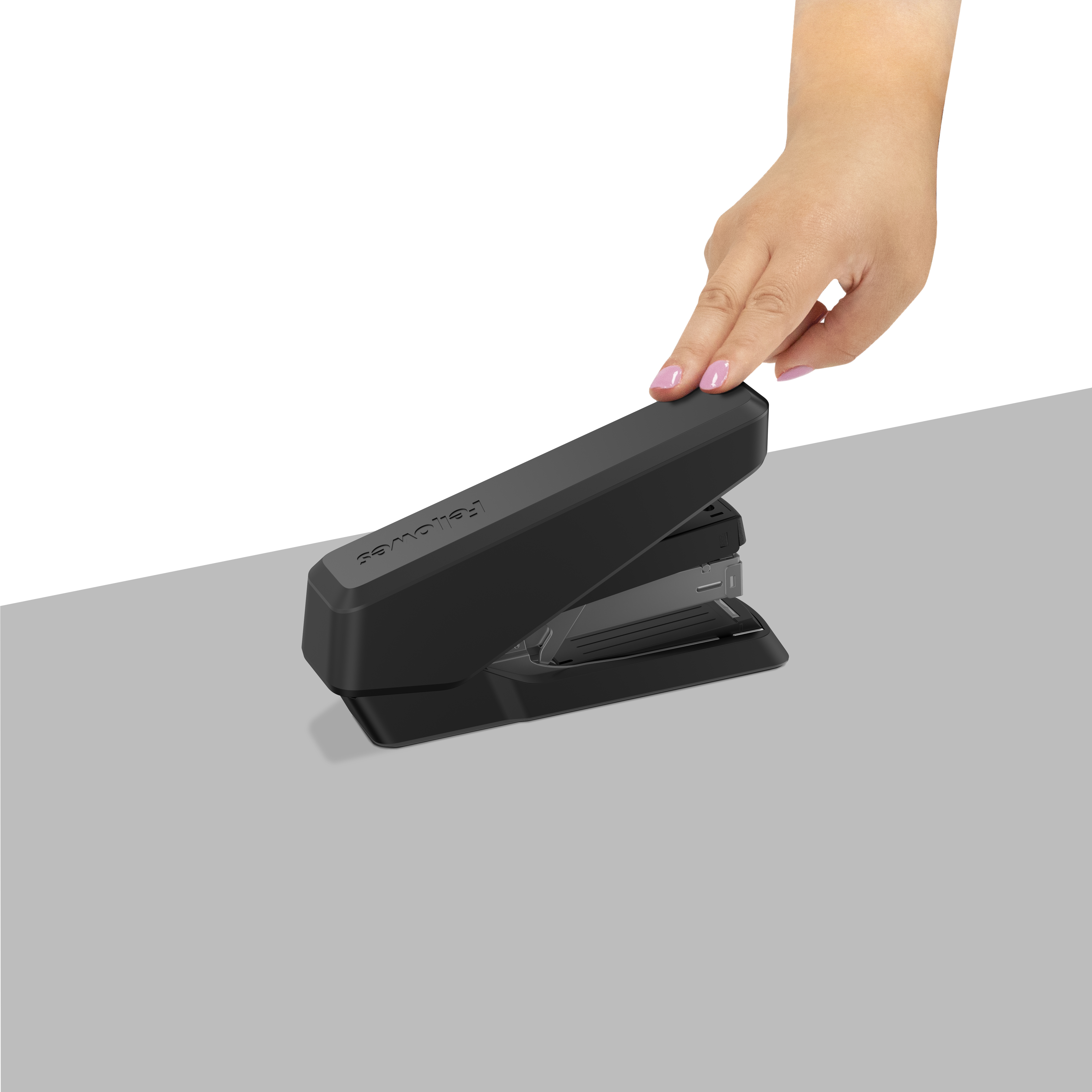 Eine Hand drückt auf einen schwarzen Tacker auf einer grauen Oberfläche, weißer Hintergrund. Rosa Nagellack sichtbar.