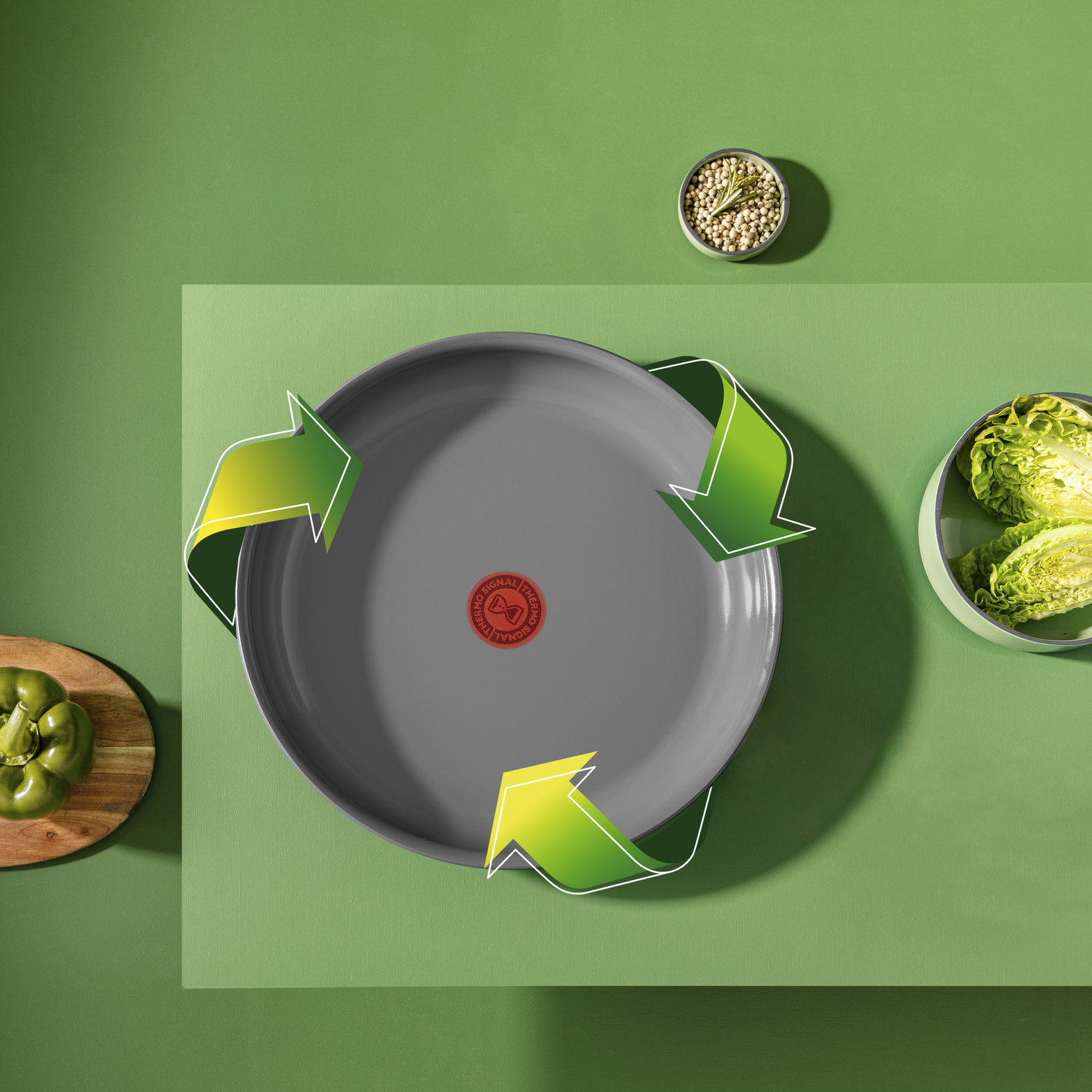 Eine graue Pfanne mit grünen Pfeilen, umgeben von grünem Paprika, einer Schüssel mit Gewürzen und Salat auf grünem Hintergrund.