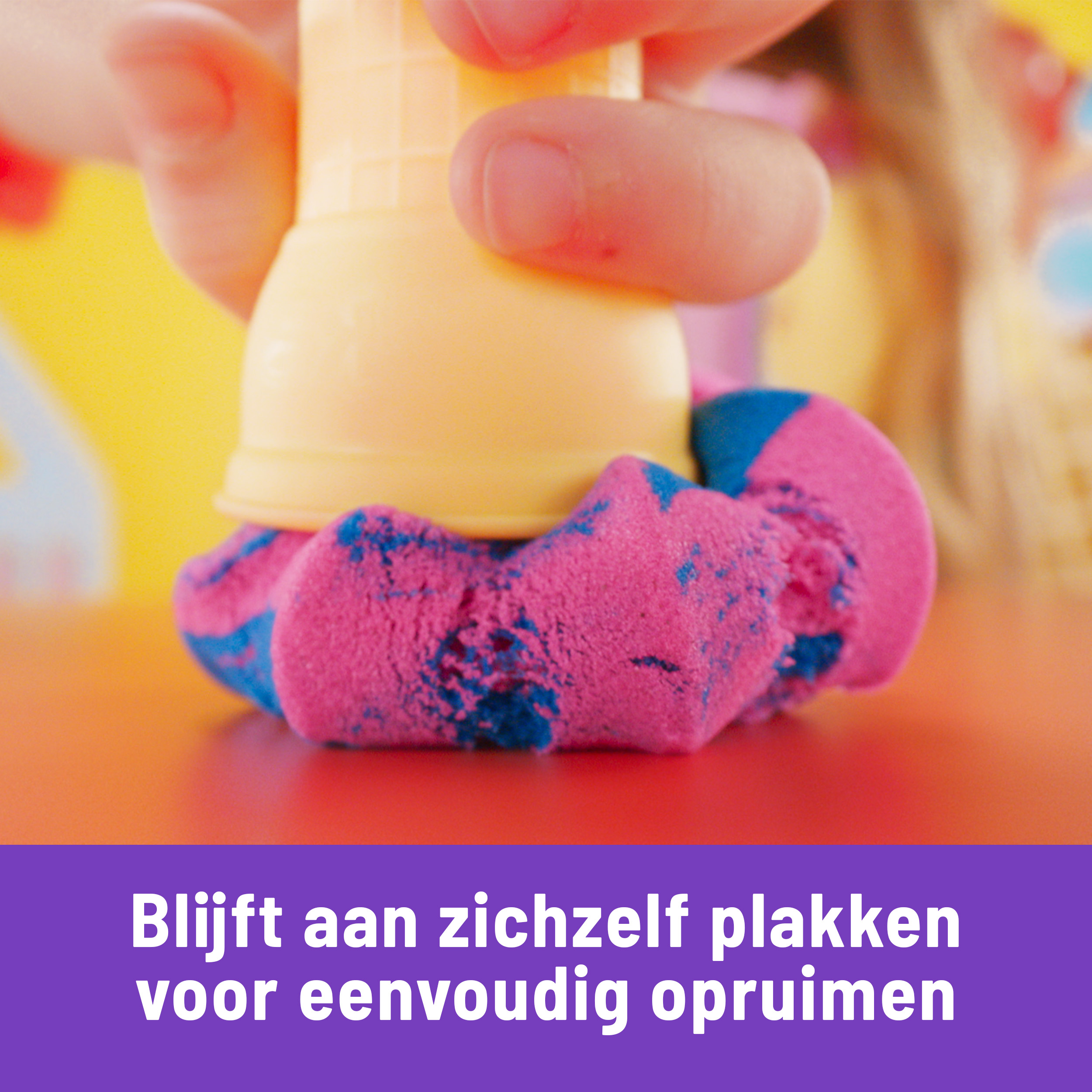 Eine Hand drückt einen gelben Kegel auf formbaren Sand in Pink und Blau, vor einem orange-gelben Hintergrund.
