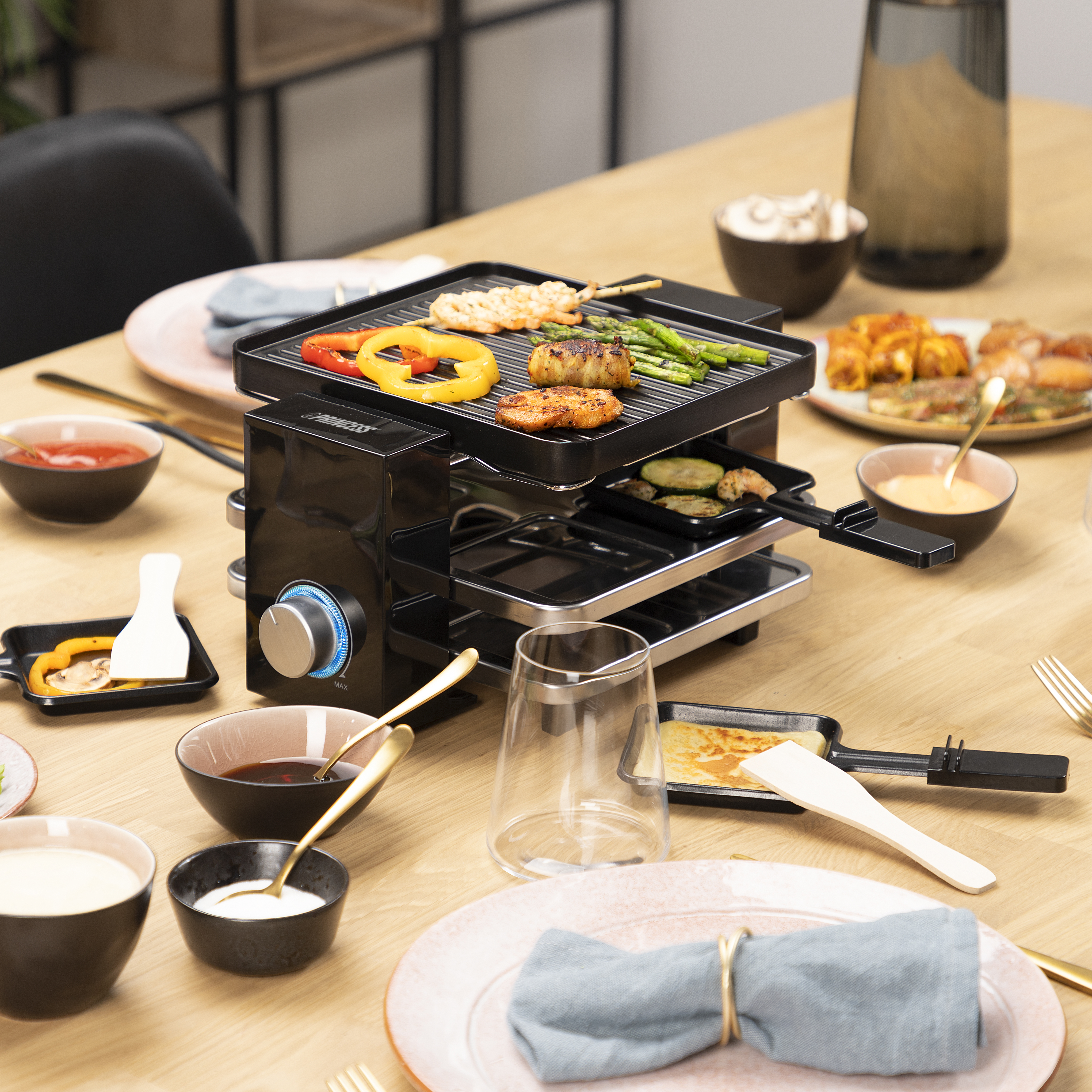 Stół z grillem do raclette, jedzeniem i talerzami. Na grillu warzywa i mięso z grilla.