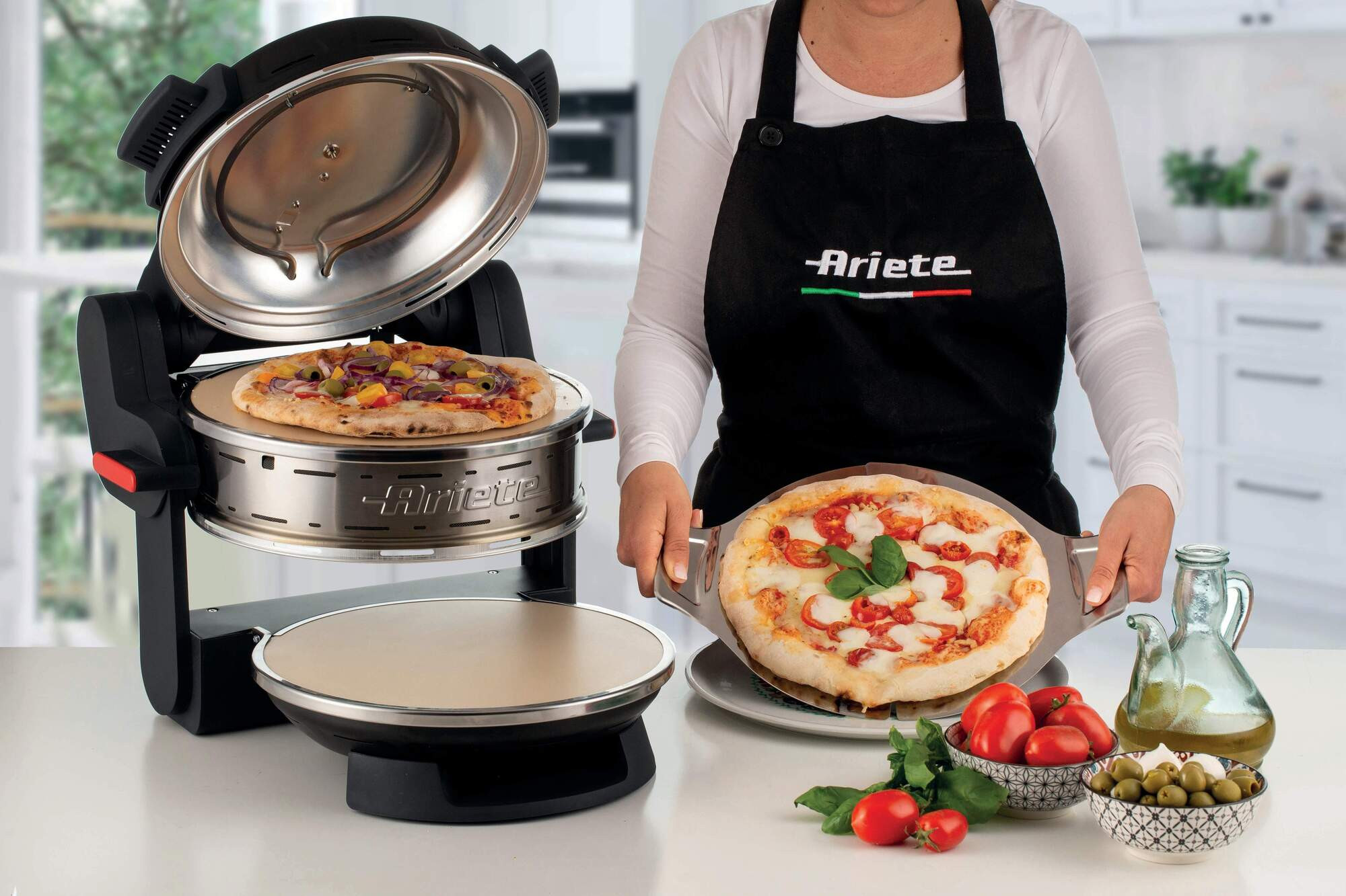Een vrouw houdt pizza vast. Een pizza-oven met een gebakken pizza erin. Tomaten, olijven en olie staan op tafel.