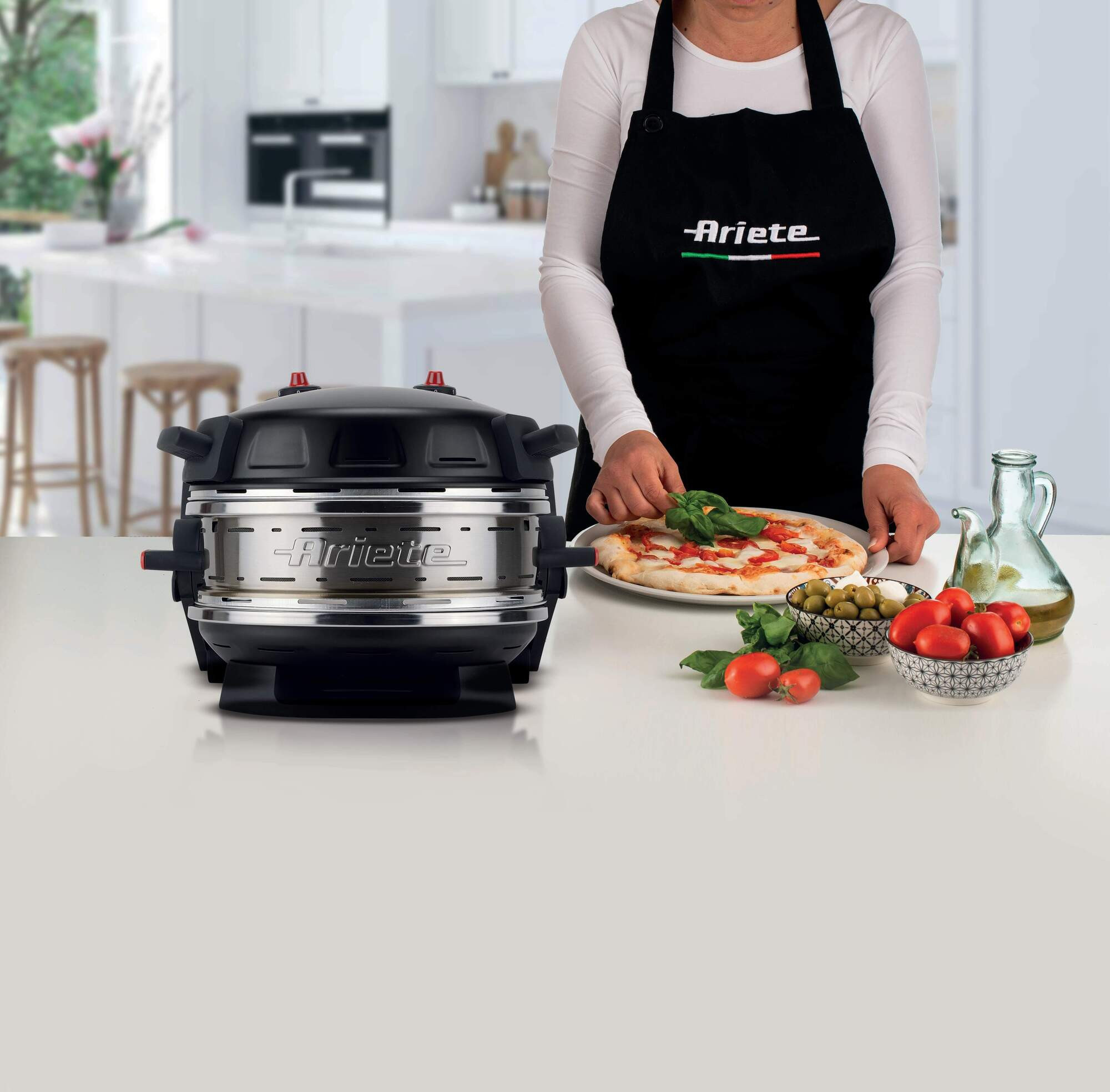 Een vrouw met een zwart schort voegt basilicum toe aan een pizza. Ingrediënten, olijfolie en een pizzaoven op tafel.
