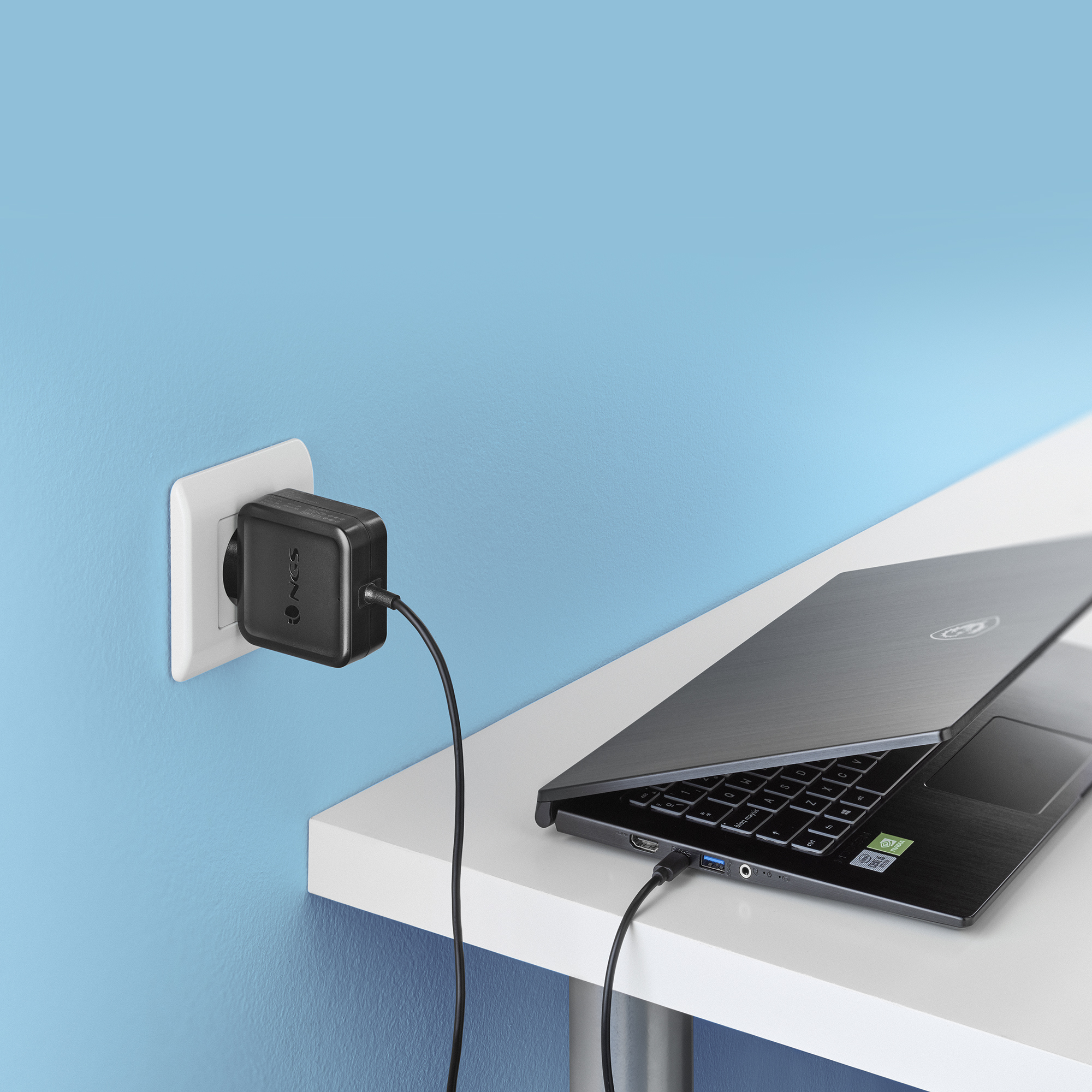 Laptop wird aufgeladen. Schwarzer Laptop auf weißem Tisch. Steckdose und Ladegerät. Blaue Wand im Hintergrund.