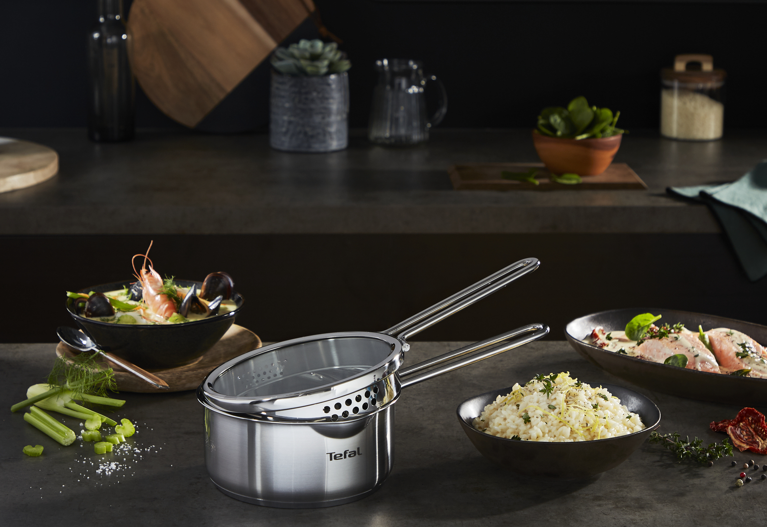 Pot Tefal en acier inoxydable avec une passoire, des bols de nourriture et des ingrédients sur un comptoir sombre. Le fond comprend des plantes et des ustensiles.