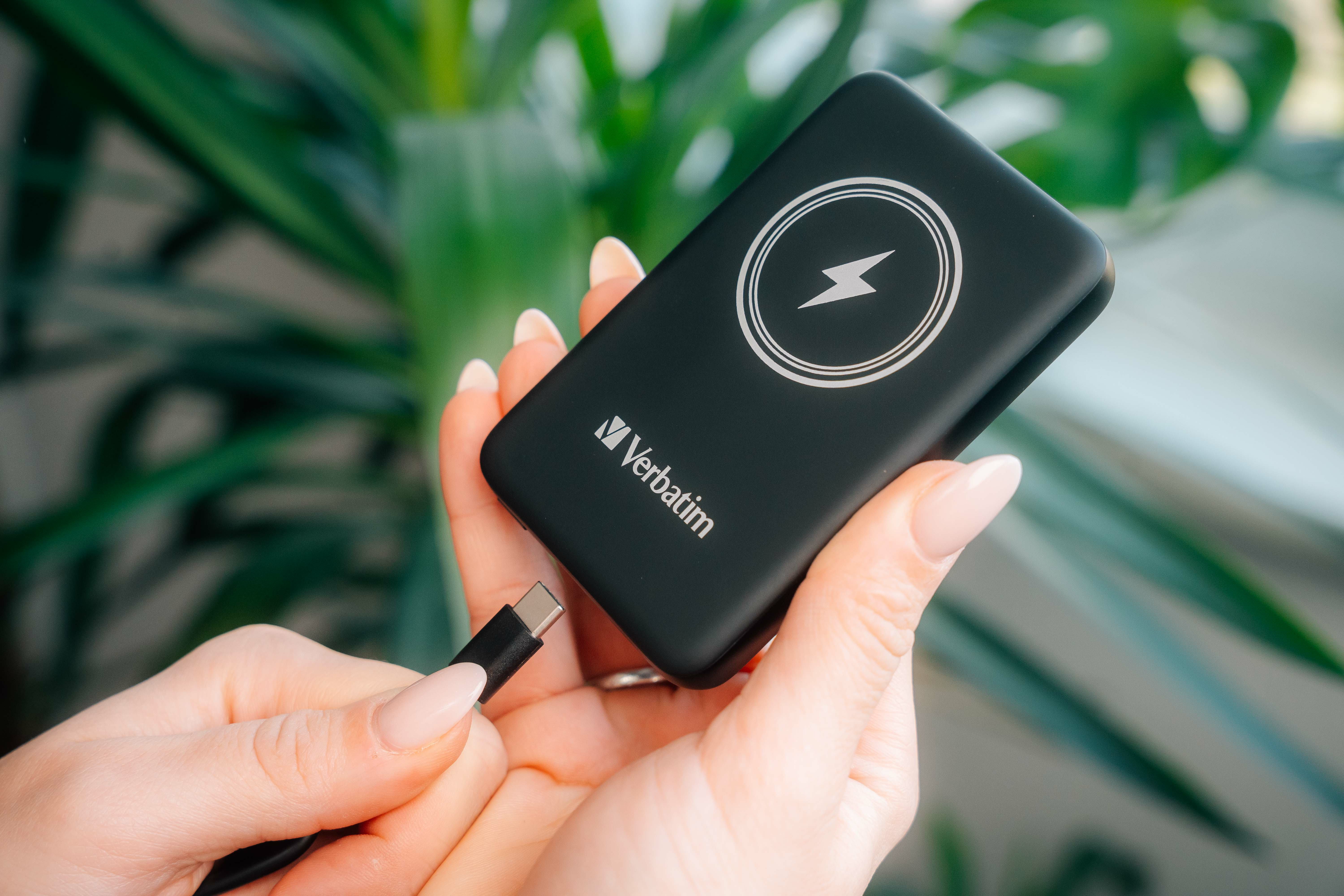 Een hand houdt een zwarte Verbatim powerbank en een USB-C-kabel vast. De achtergrond is een wazige groene plant.