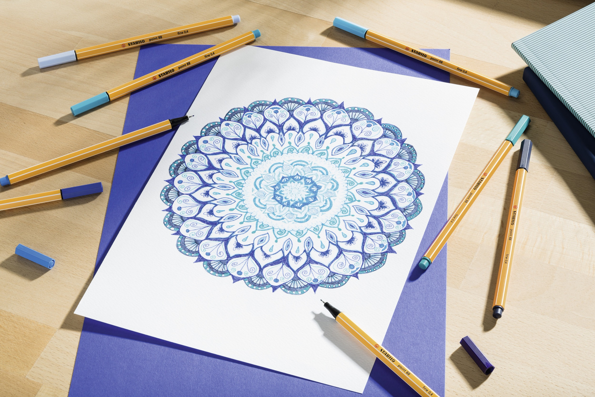 Un disegno di mandala su carta bianca, circondato da penne colorate. Lo sfondo ha carta blu e una superficie in legno.