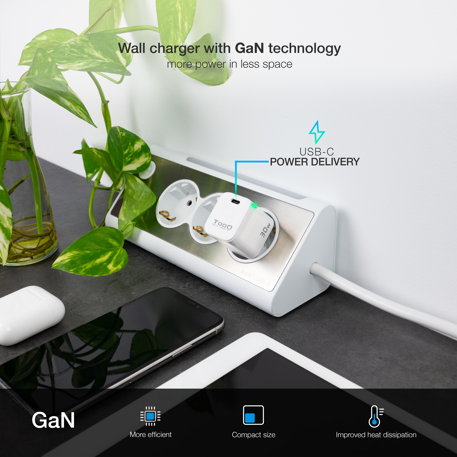 Una regleta blanca con un cargador USB-C está enchufada. Plantas verdes con hojas en el fondo.