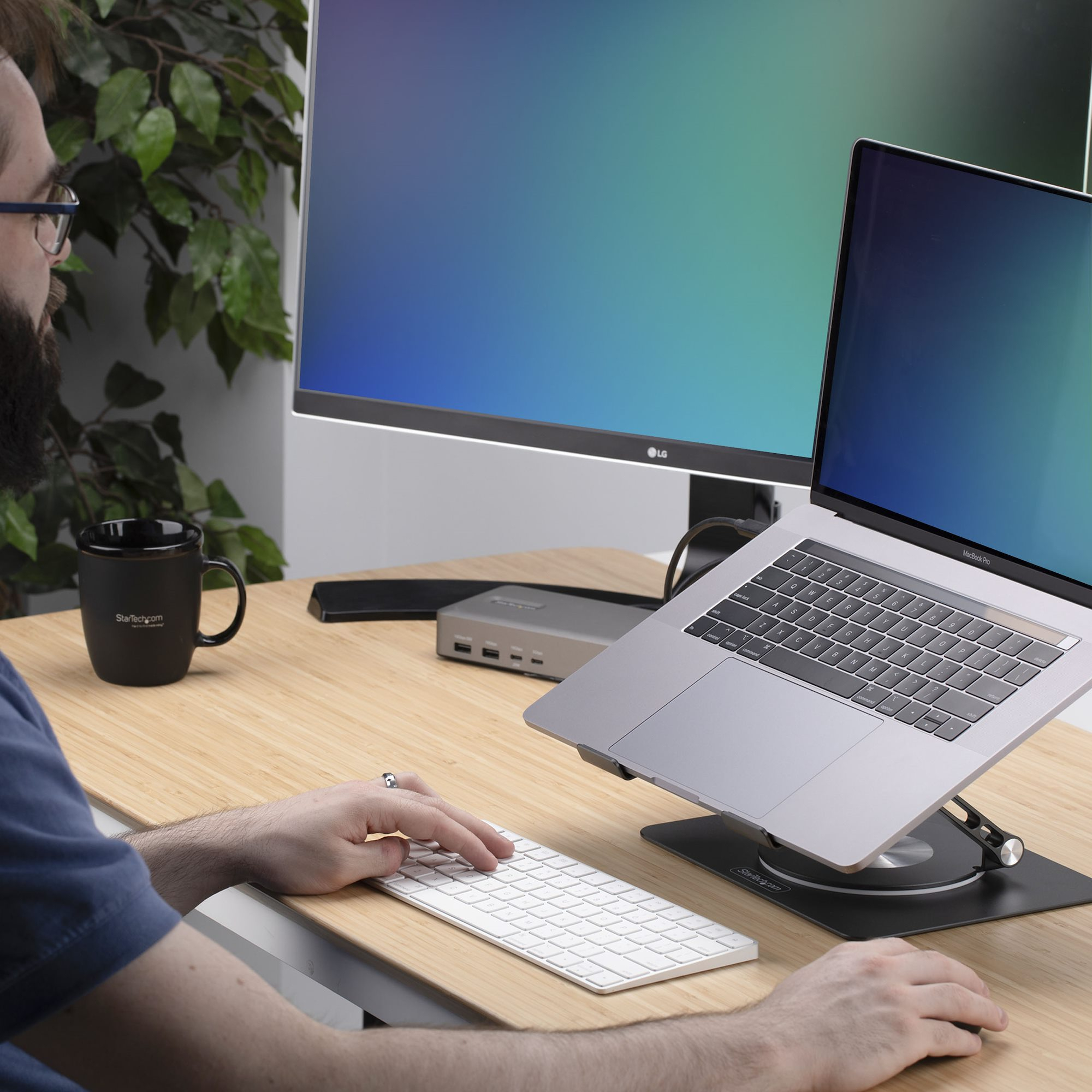 Ein Mann benutzt einen Laptop auf einem Ständer mit Tastatur und Maus. Ein Monitor und eine Tasse sind ebenfalls auf dem Schreibtisch.