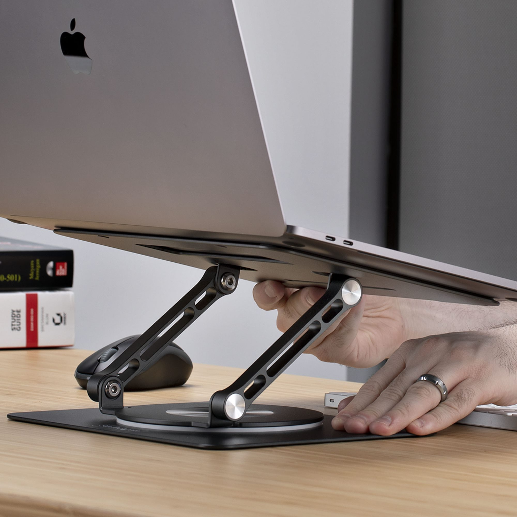 Laptop auf schwarzem Ständer, Höhenverstellung. Maus und Bücher liegen auf dem Schreibtisch. Eine Hand stellt den Ständer ein.