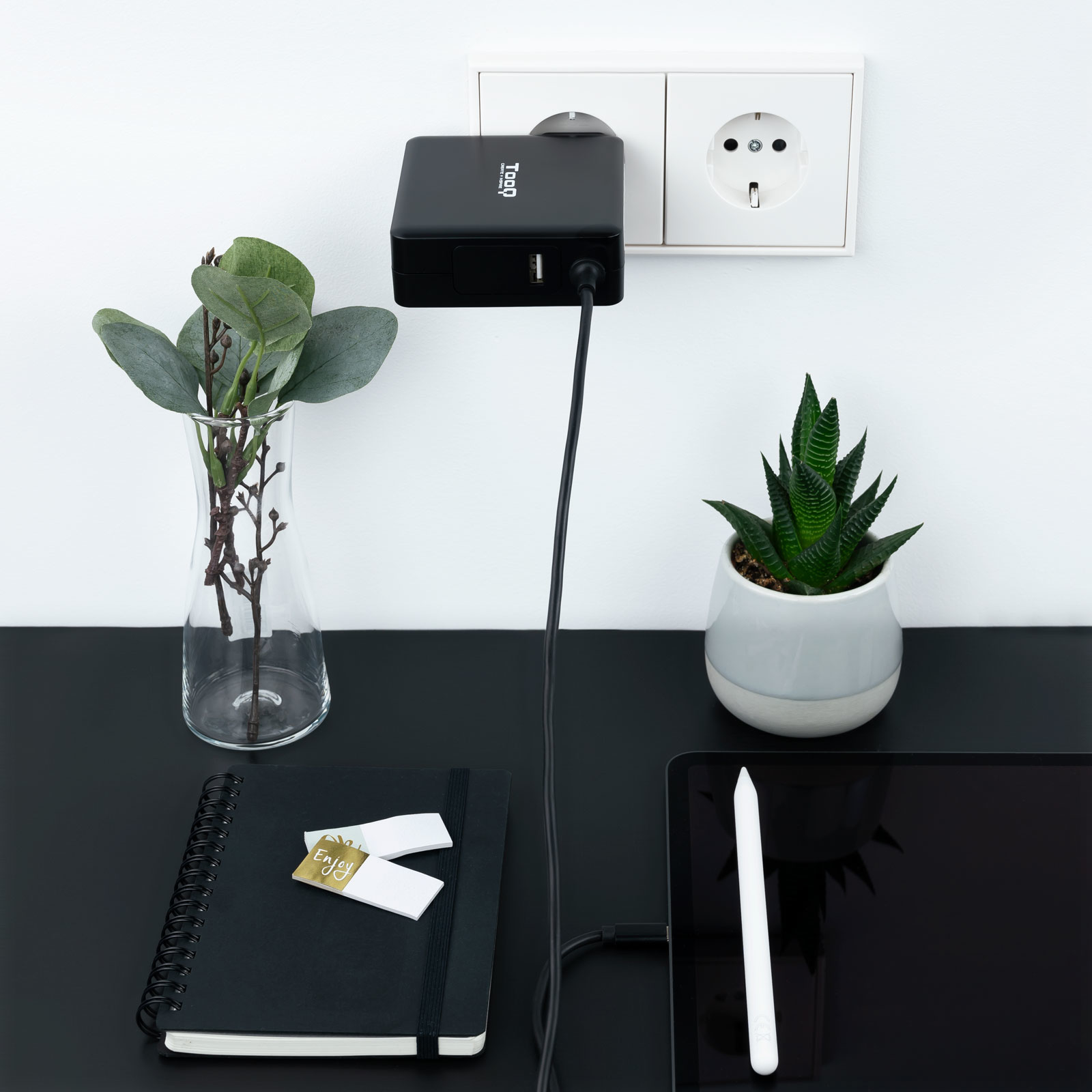 Cargador negro enchufado, cable conectado a una tableta. Plantas y cuaderno en el escritorio.