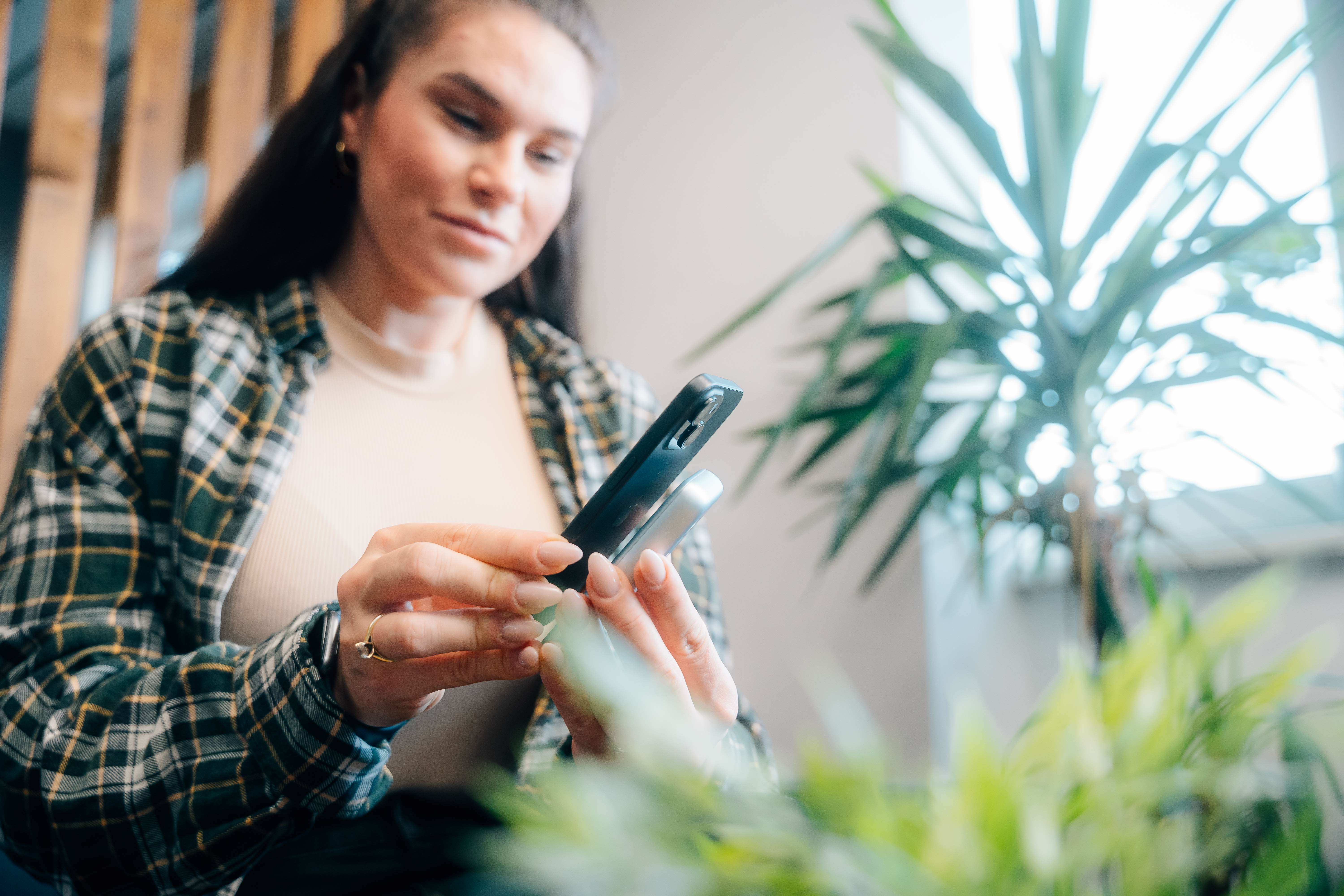 Een vrouw houdt twee telefoons vast. Ze draagt een geruit overhemd. Een plant staat op de achtergrond.