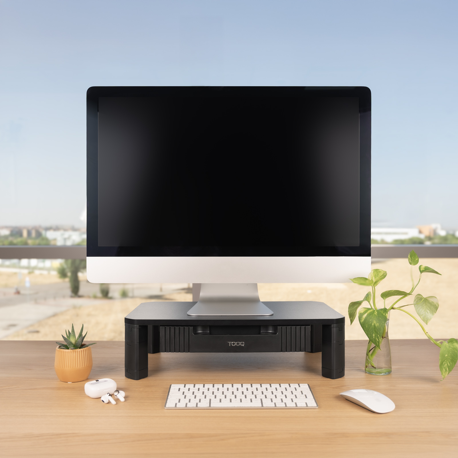 Un monitor per computer è posizionato su un supporto nero su una scrivania di legno. Sono presenti anche una tastiera, un mouse e una pianta.