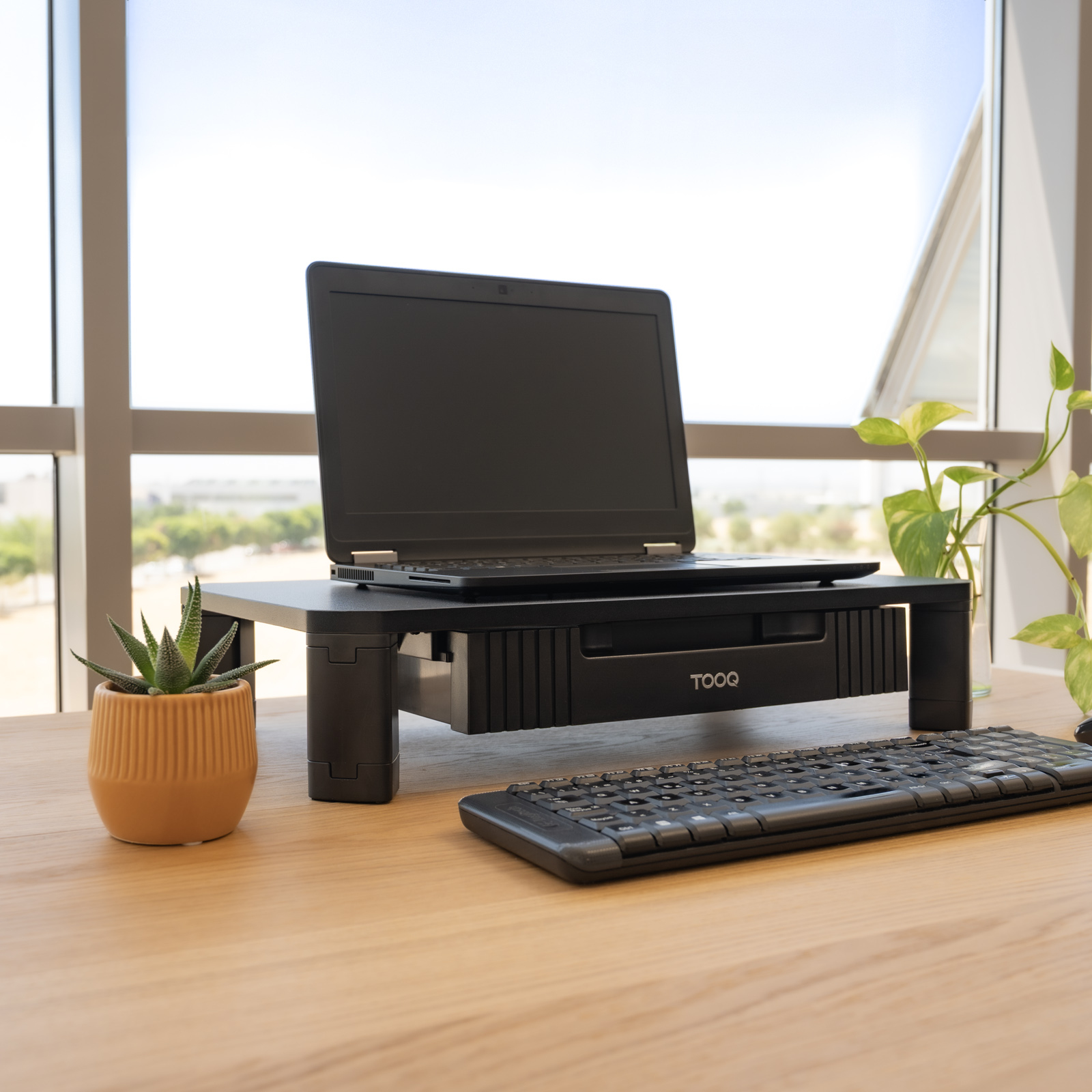 Un laptop su un supporto nero con tastiera, una pianta e una succulenta su una scrivania di legno.