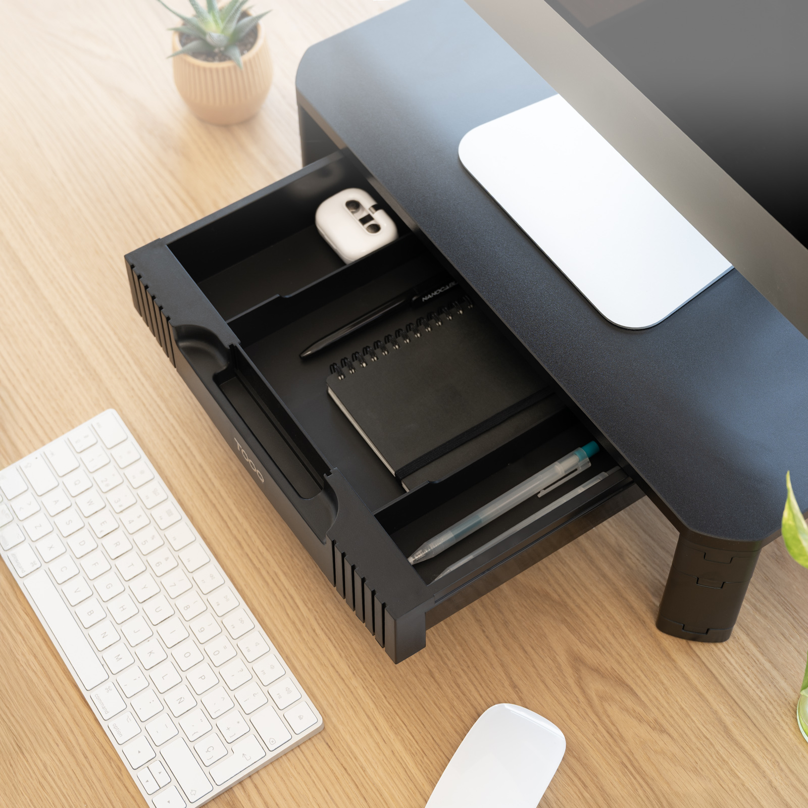 Una scrivania con supporto per monitor, tastiera e mouse. Un cassetto contiene un quaderno, una penna e un accessorio bianco.