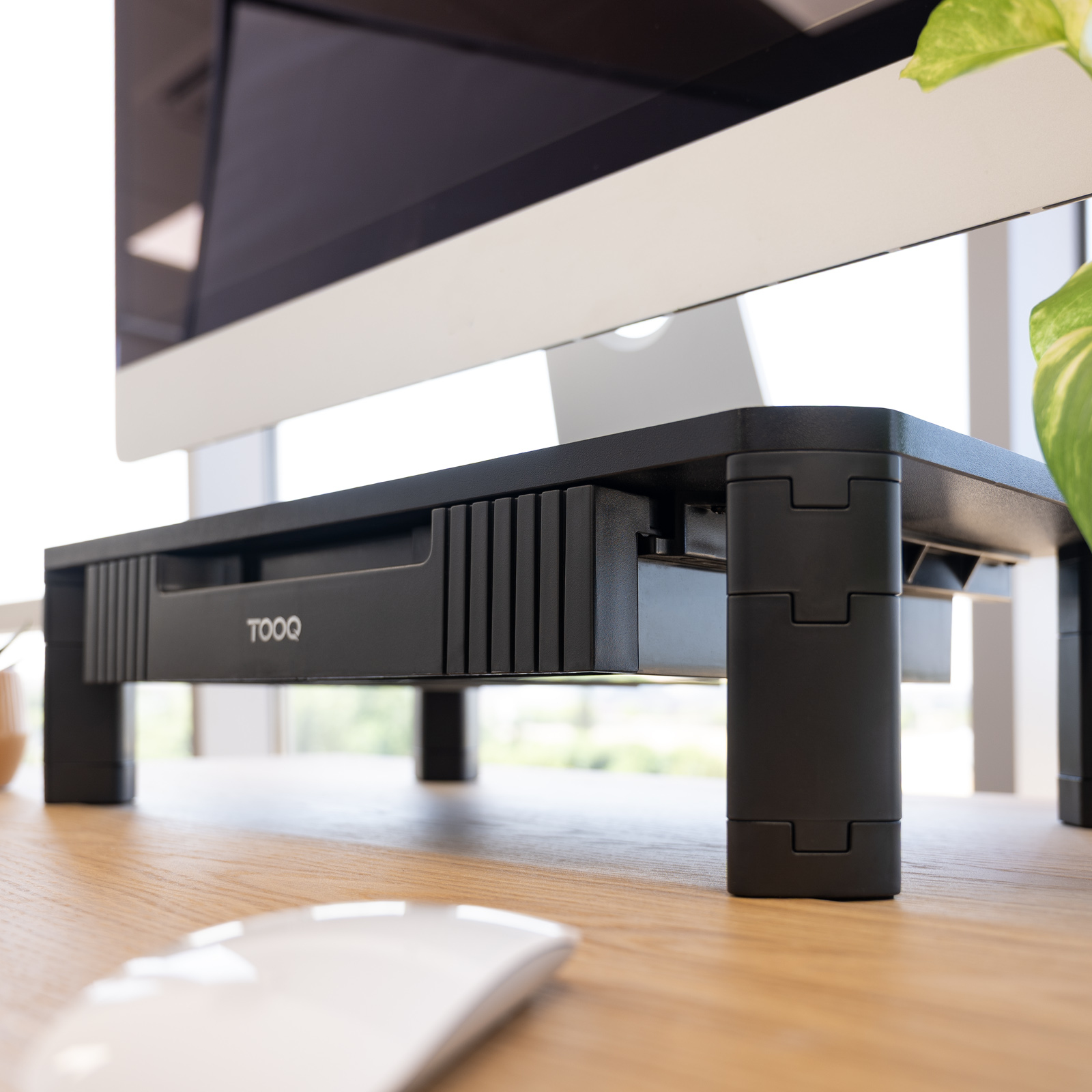 Un supporto per monitor nero con cassetto, un mouse bianco su una scrivania di legno. Uno schermo di computer sullo sfondo.