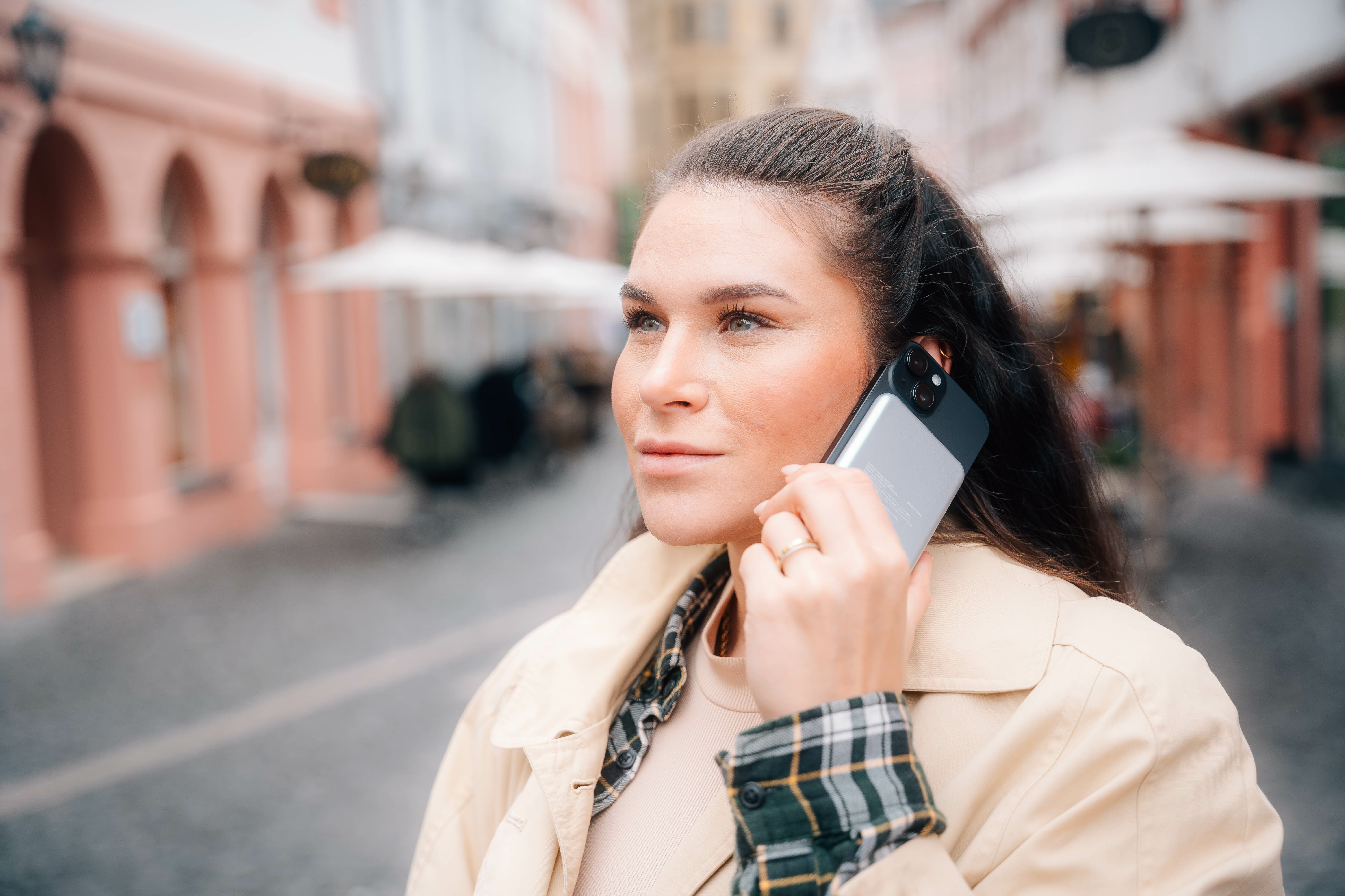 Vrouw aan de telefoon op straat. Ze draagt een beige jas en een geruit overhemd. Gebouwen op de achtergrond.