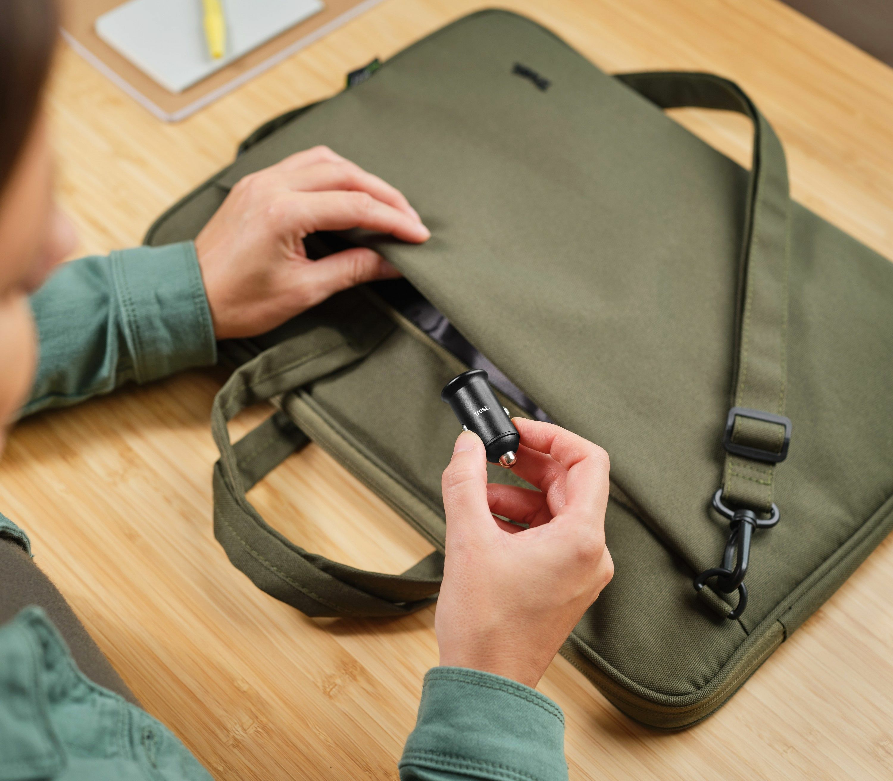 Una persona sostiene un cargador de coche negro junto a una funda para portátil verde oliva.