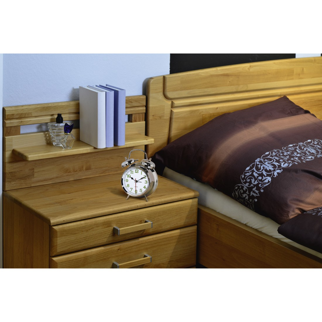 Una escena de dormitorio con una mesita de noche de madera, despertador, libros y una cama con edredón marrón.