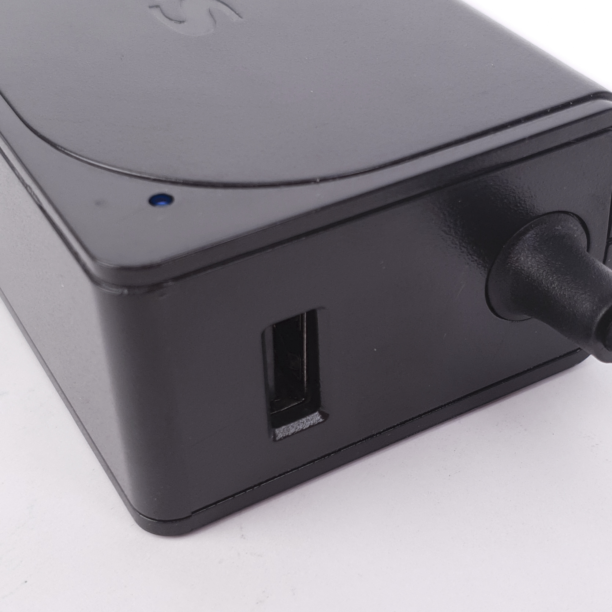 Primer plano de un adaptador de corriente negro con puerto USB. Se ve una luz azul. El fondo es blanco.