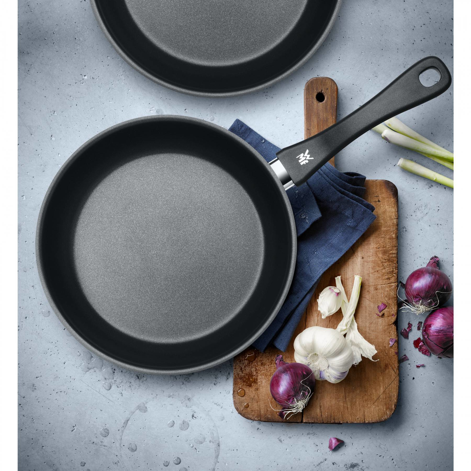 Zwei Pfannen, Holzbrett mit Zwiebeln und Knoblauch sowie Frühlingszwiebeln. Grauer Hintergrund.
