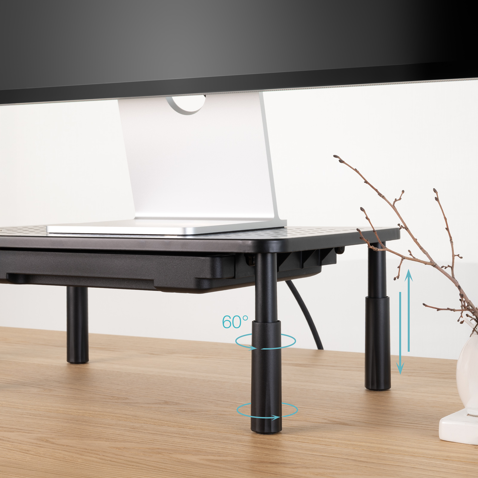 Un laptop è su un supporto per monitor nero con gambe regolabili. Un vaso con rami è su una scrivania di legno.