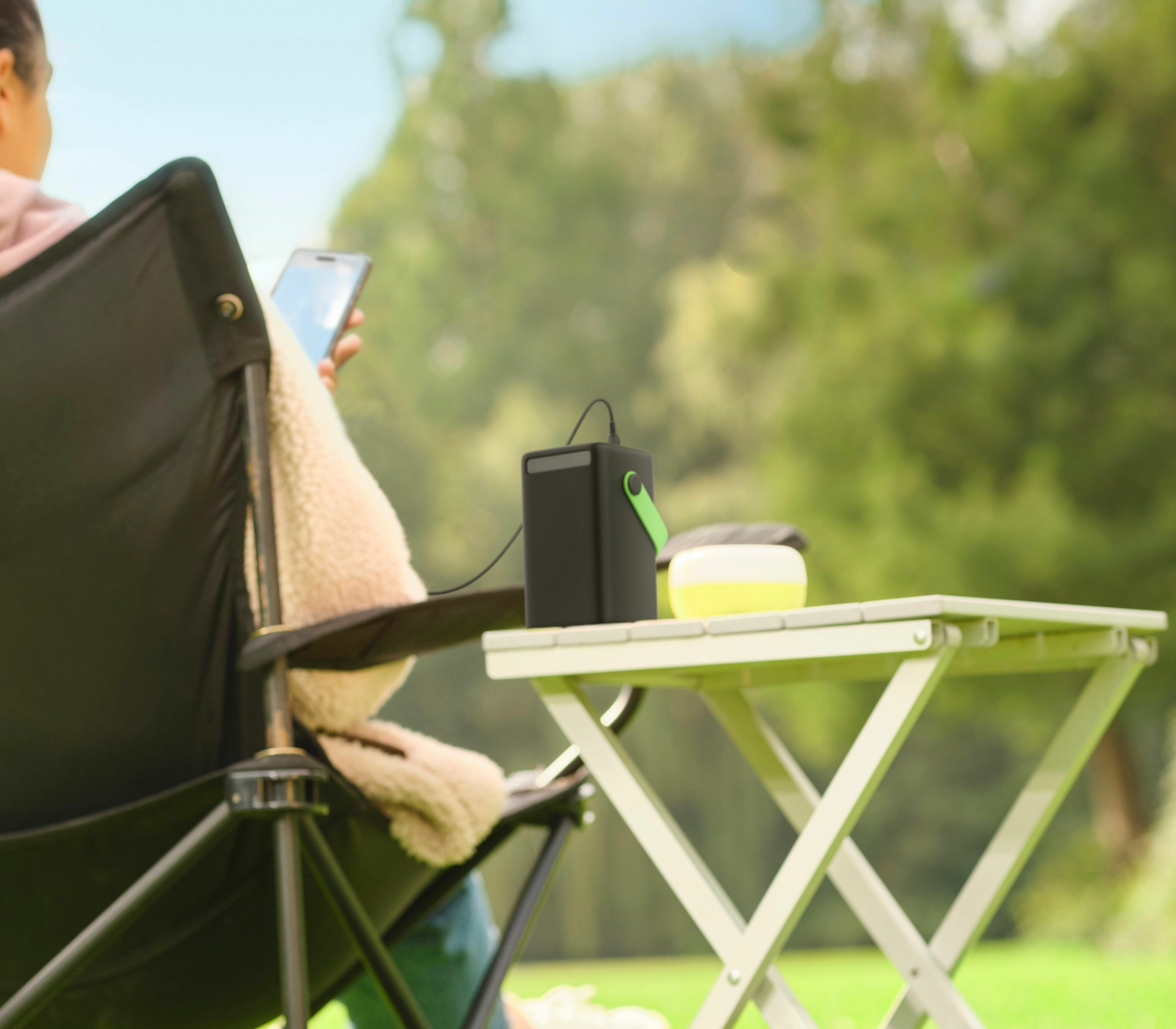 Person im Stuhl, die ein Telefon benutzt, neben einem Tisch mit Lautsprecher und Lampe auf einer grünen Wiese.