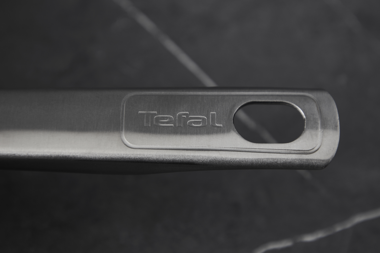 Nahaufnahme eines Edelstahlpfannengriffs mit dem 'Tefal'-Logo und einem Aufhängeloch, vor dunklem Hintergrund.