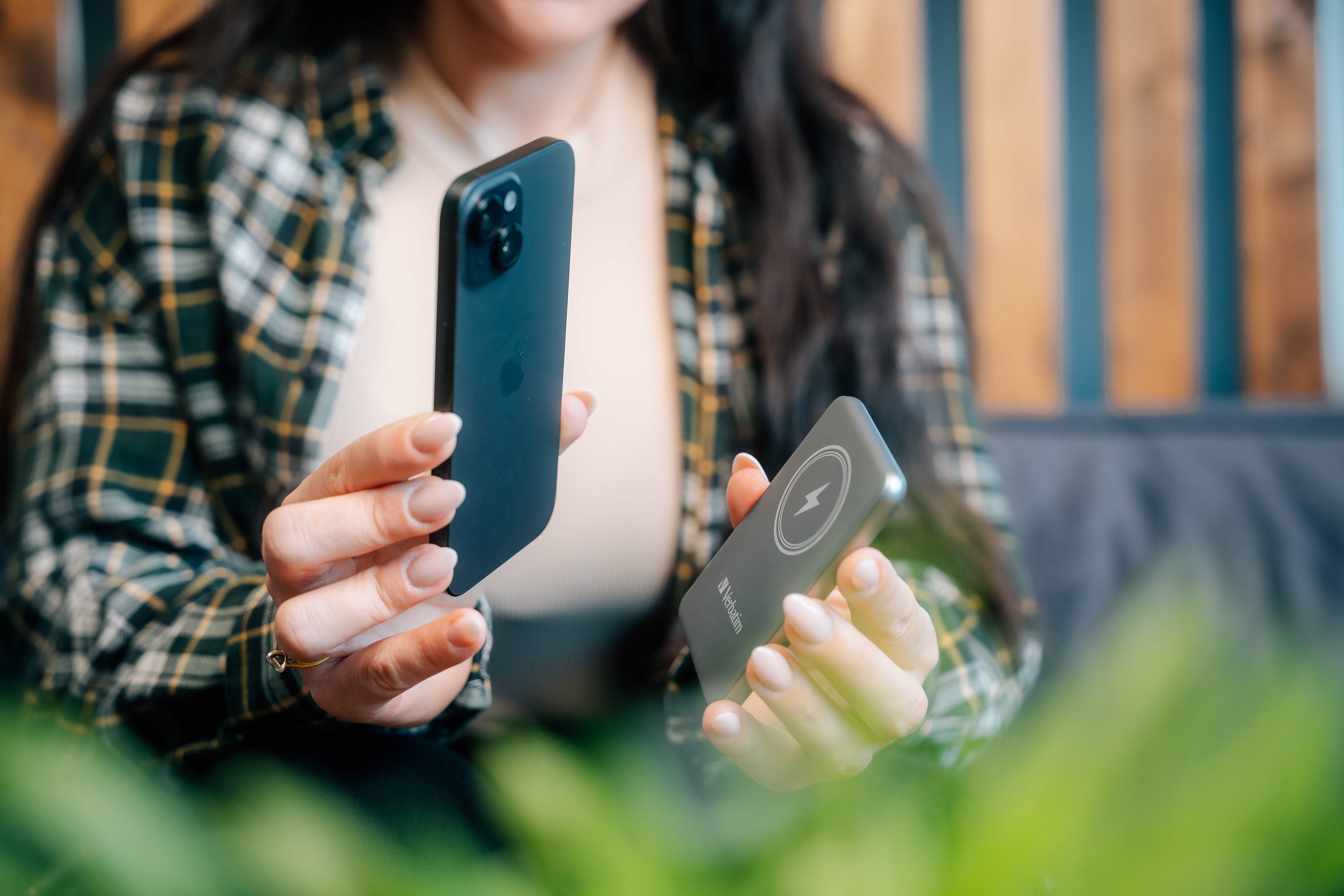 Een persoon houdt een zwarte telefoon en een oplader vast. De persoon draagt een geruit overhemd. Groene planten op de voorgrond.