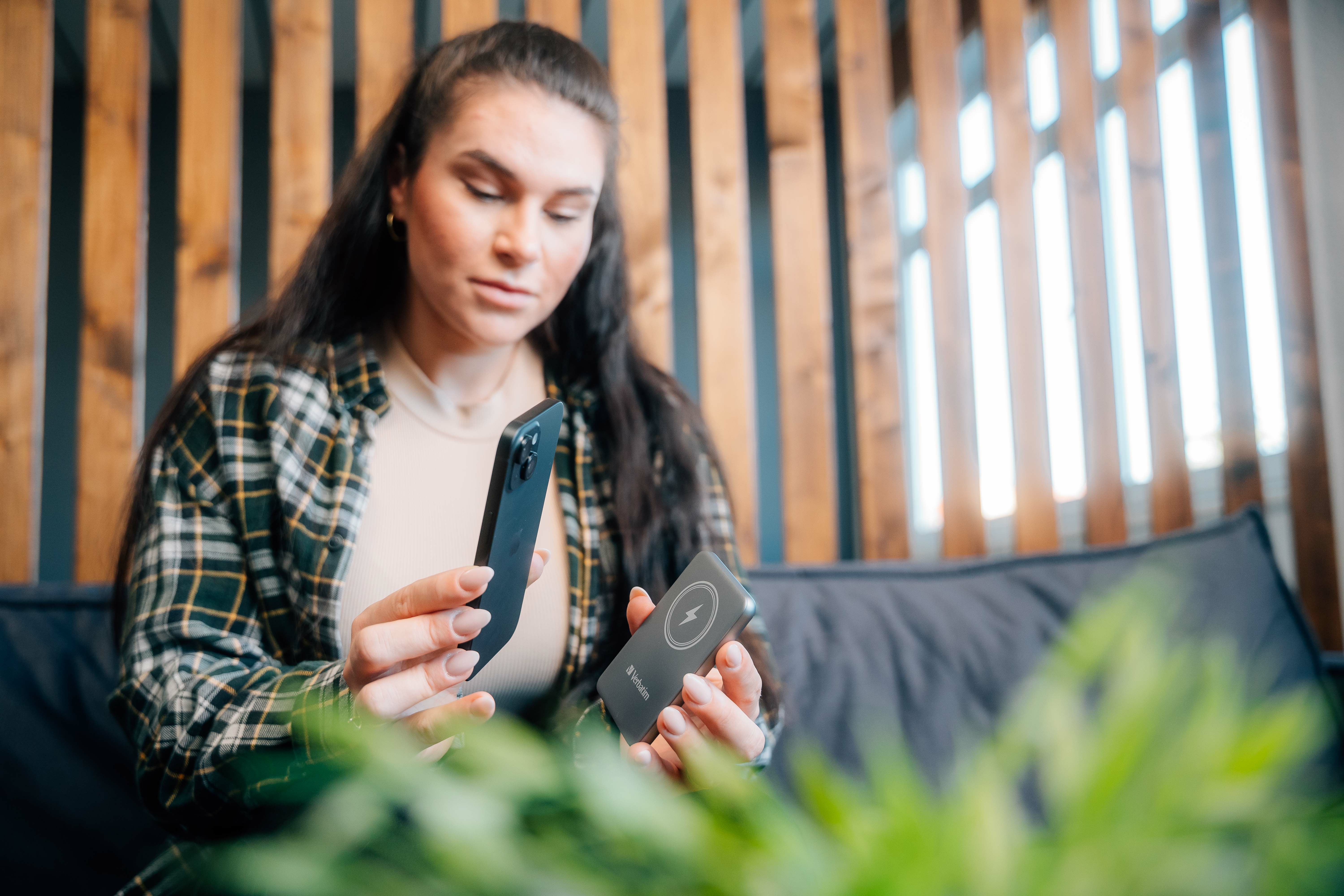 Een vrouw houdt een telefoon en een oplader vast. Ze draagt een geruit overhemd. Achtergrond heeft houten latten en een bank.