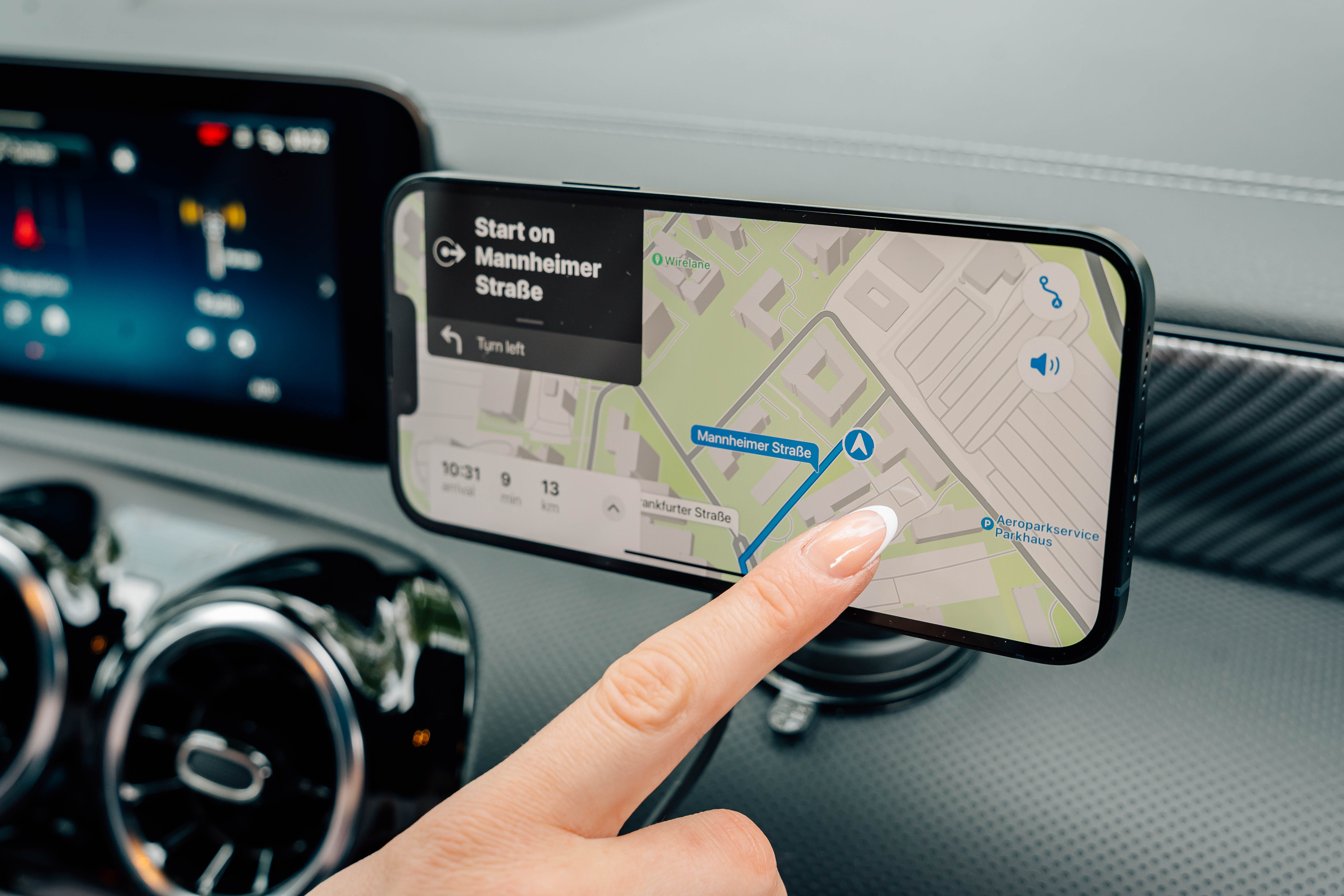 Eine Hand berührt ein Telefon, das eine Karte mit Navigation anzeigt. Der Bildschirm zeigt 'Mannheimer Straße' und 'Turn left'.
