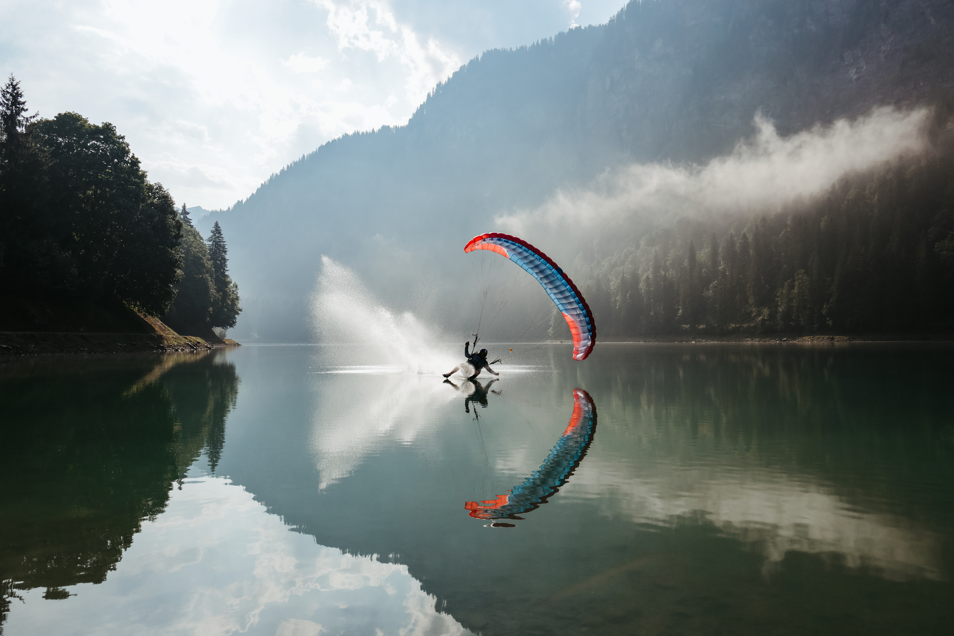 A paraglider glides over a lake, with mountains and trees in the background. The paraglider's reflection is visible.