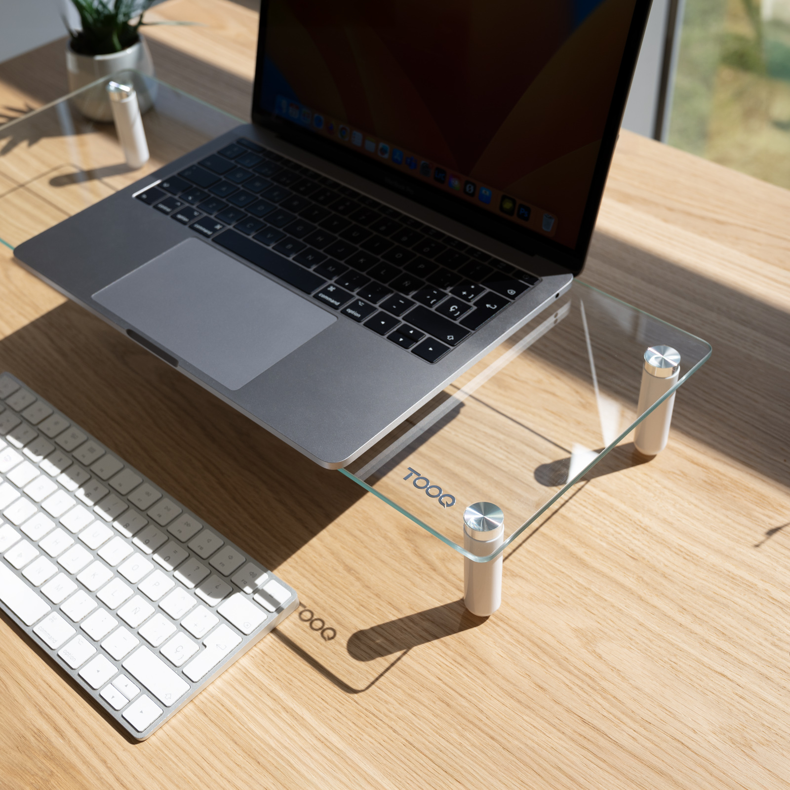 Laptop auf einem Glasständer über einem Holztisch, Tastatur links. Eine Pflanze befindet sich im Hintergrund.