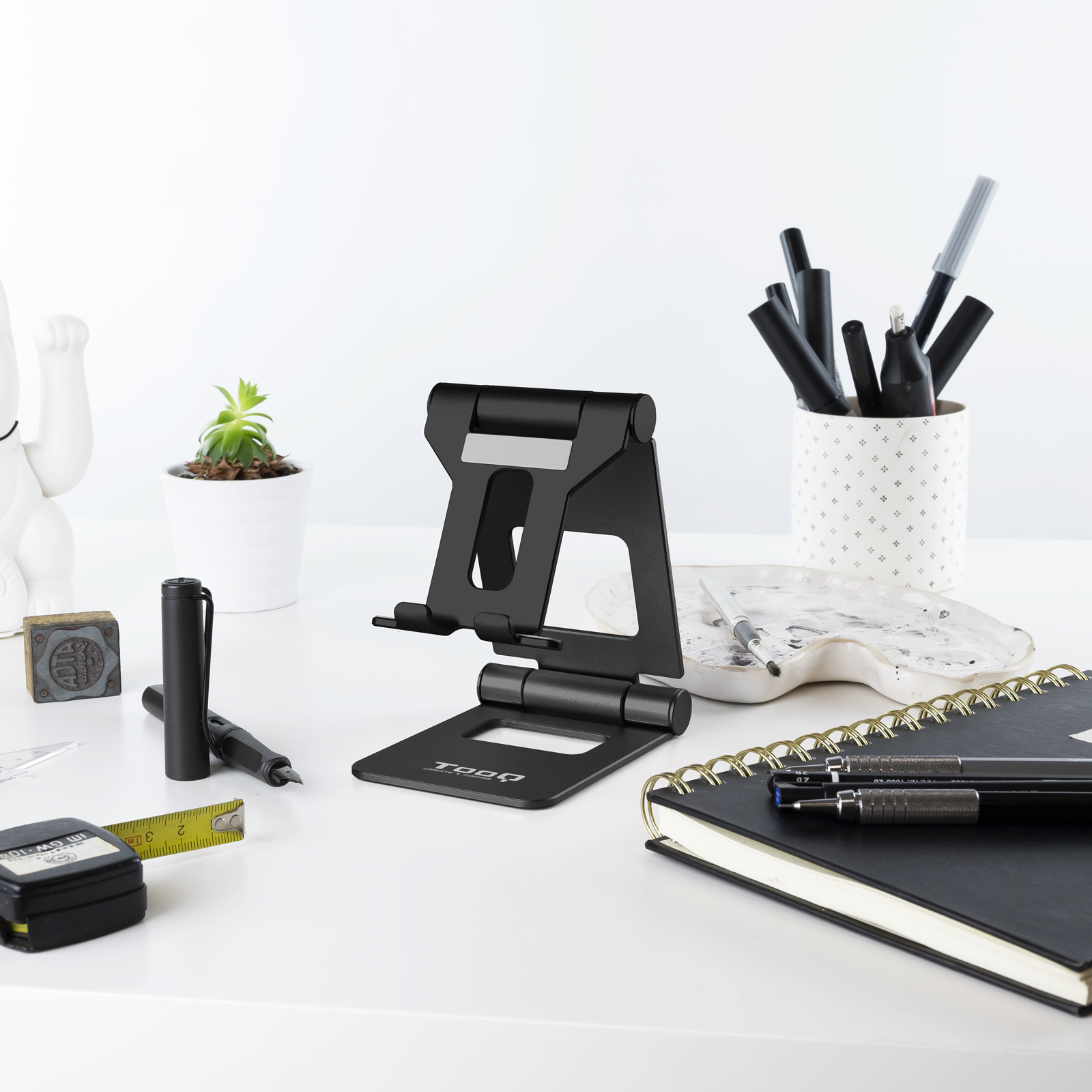 Soporte para teléfono negro sobre escritorio blanco, bolígrafos, cuaderno, planta, cinta métrica y figura de gato blanco.