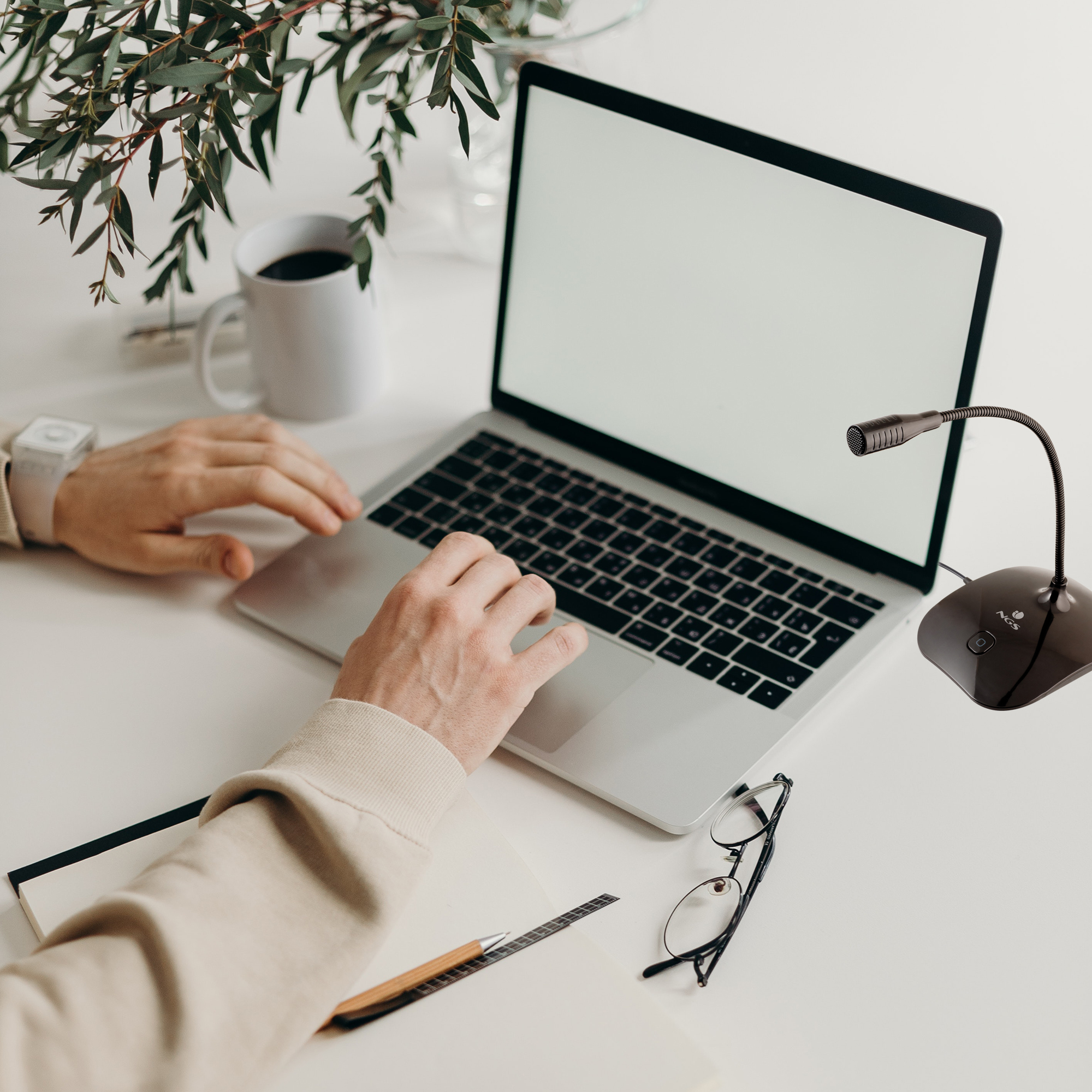 Osoba korzystająca z laptopa na białym biurku. Jest mikrofon, kubek do kawy, okulary i długopis.