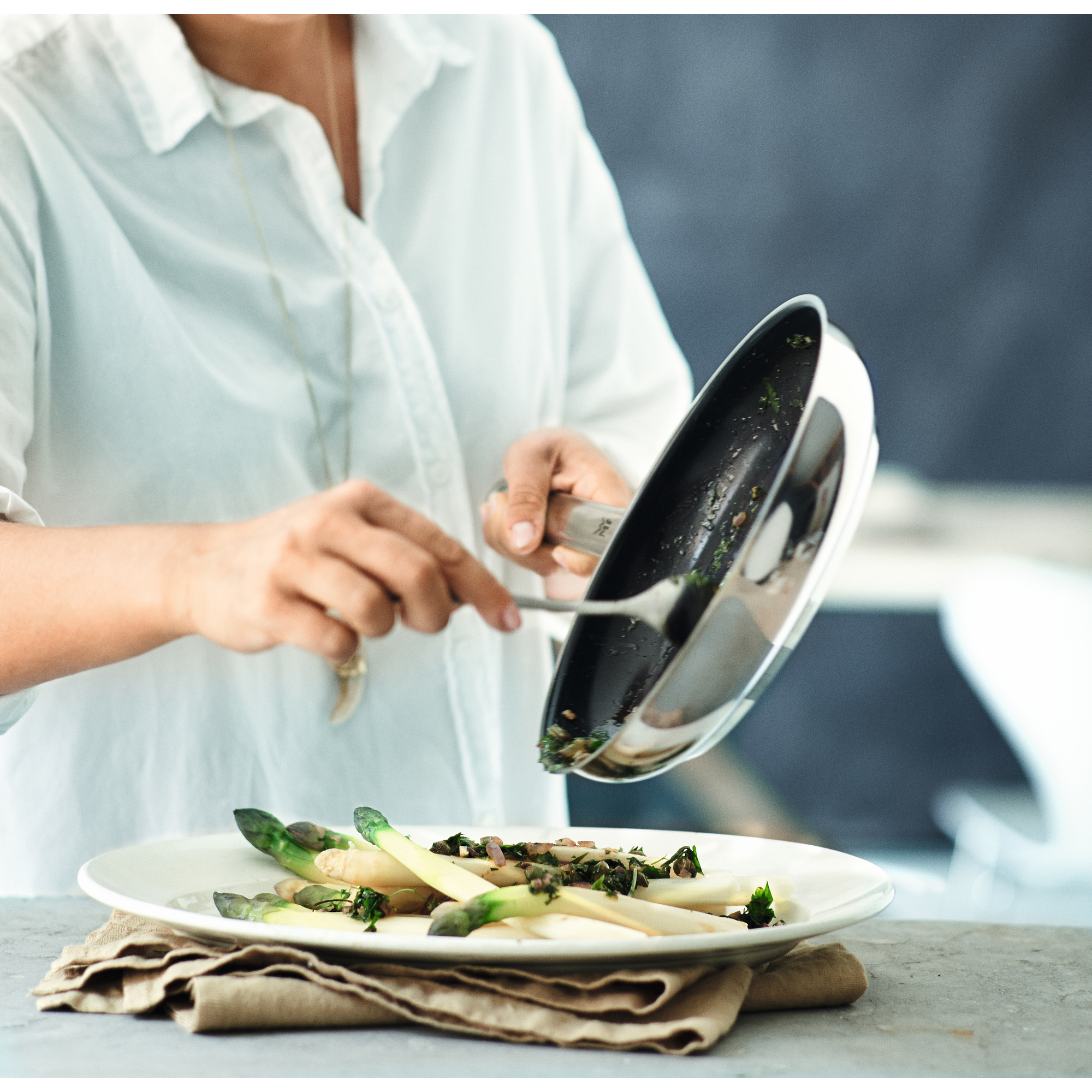 Eine Person gießt Essen aus einer Pfanne auf einen Teller Spargel. Der Teller steht auf einem Leinentuch.