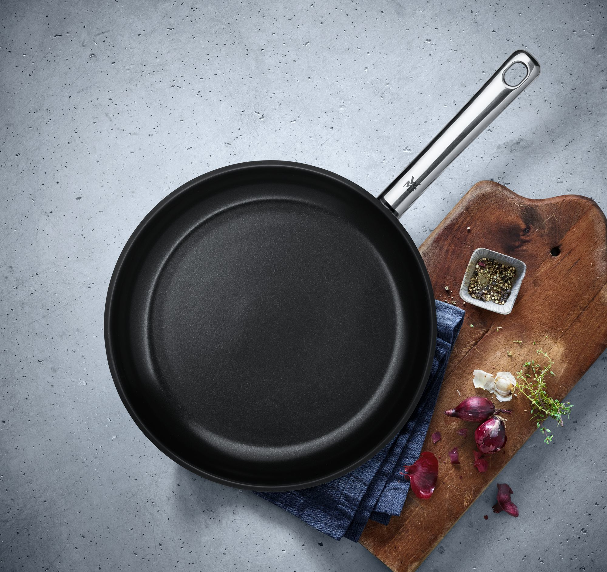 Una padella nera con manico argentato, su una superficie grigia. Vicino ci sono ingredienti come cipolle e spezie.