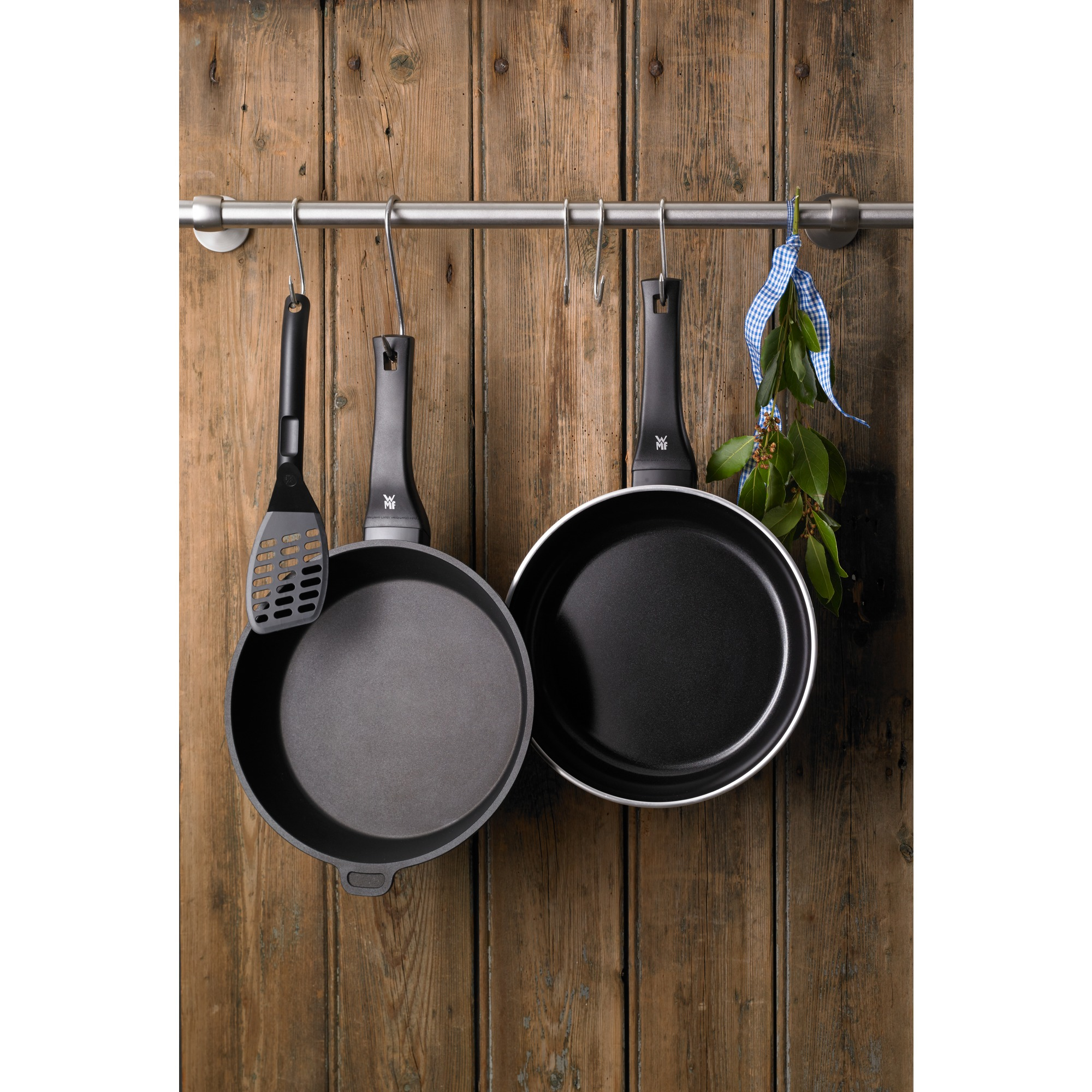 Zwei schwarze Bratpfannen und ein Pfannenwender hängen an einer Metallstange vor einer Holzwand. Kräuter mit blauem Band rechts.