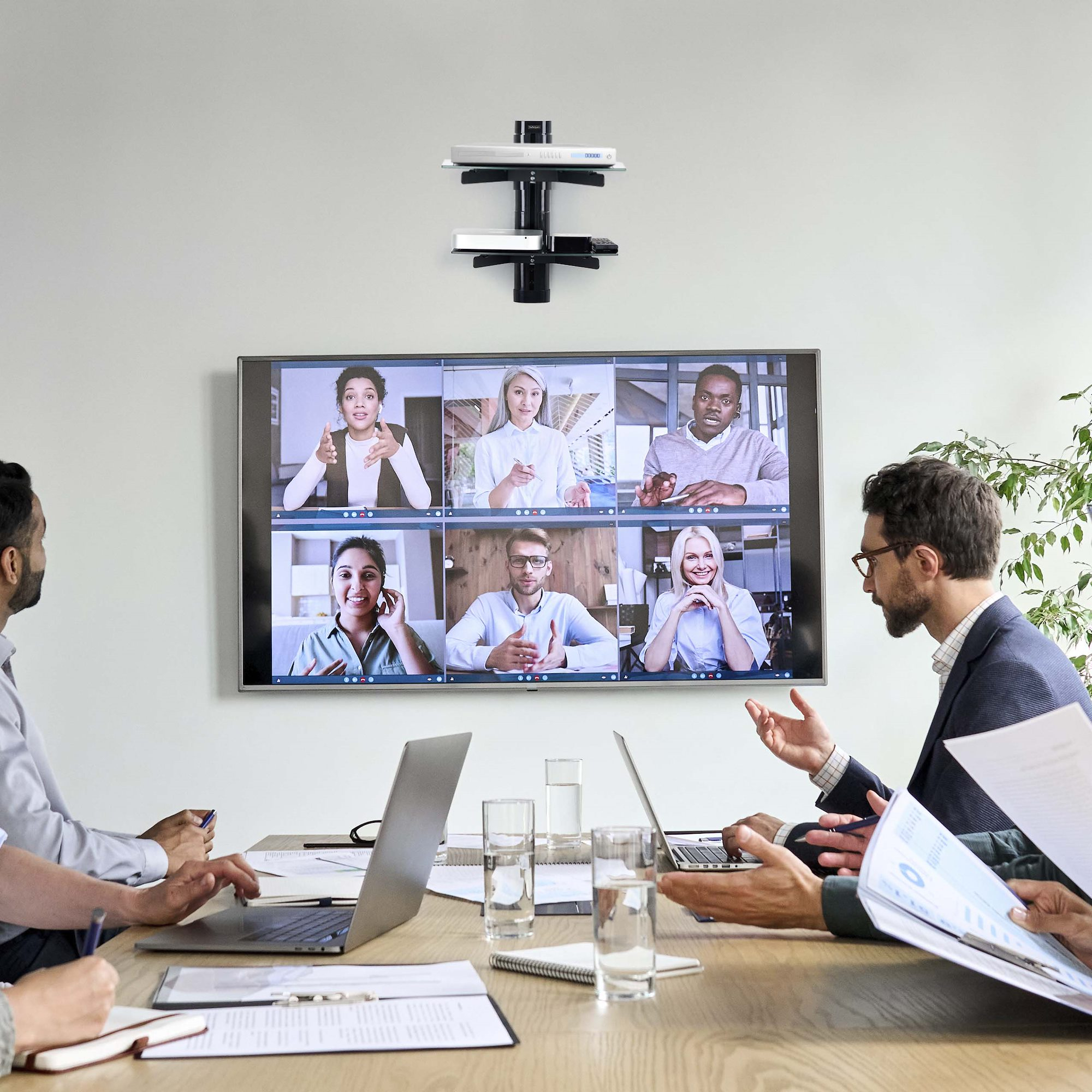 Ein Meeting mit Personen auf dem Bildschirm. Personen befinden sich in einem Konferenzraum mit Laptops, Wassergläsern und einer Pflanze.