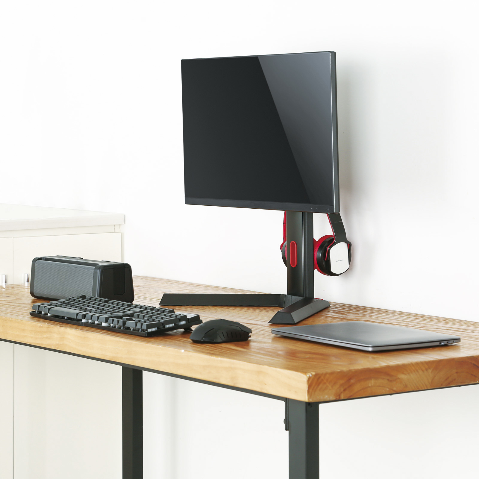 Configuración de escritorio: monitor, teclado, ratón, altavoz, auriculares y portátil sobre un escritorio de madera contra una pared blanca.